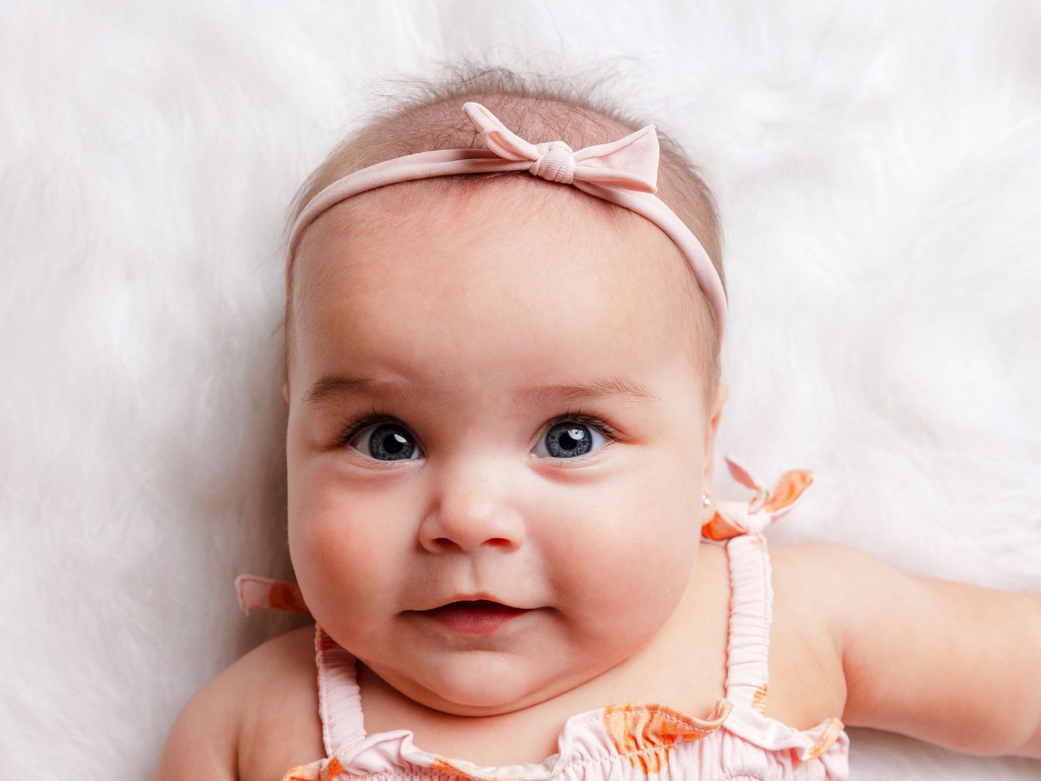 Adorable baby girl wearing a pink bow headband and floral outfit, lying on a soft white fluffy blanket.