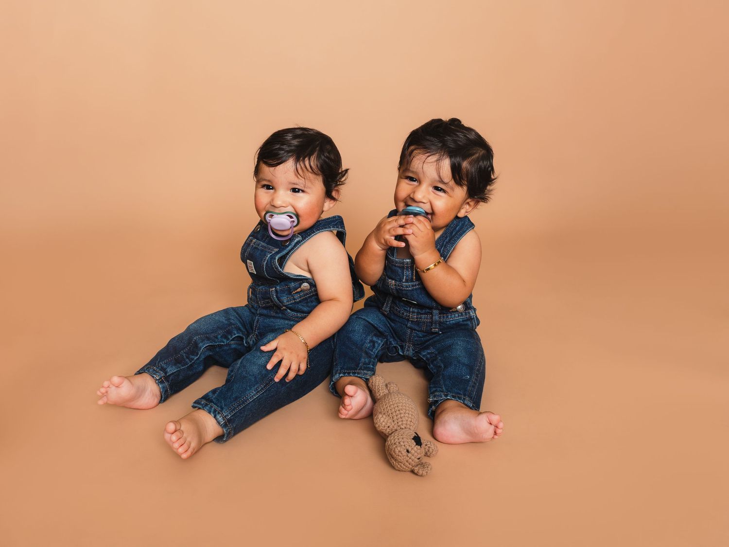Two babies wearing matching denim outfits lay on a peach-colored backdrop in a series of playful poses.