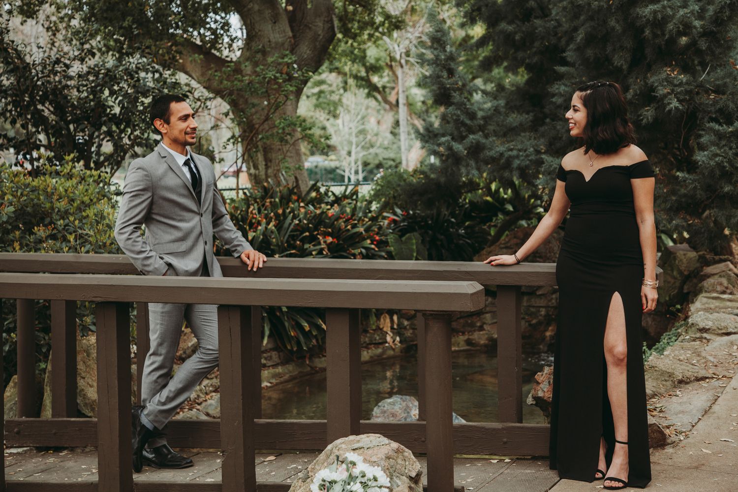 A couple in formal attire poses on a wooden bridge surrounded by lush greenery, man in gray suit, woman in black gown.