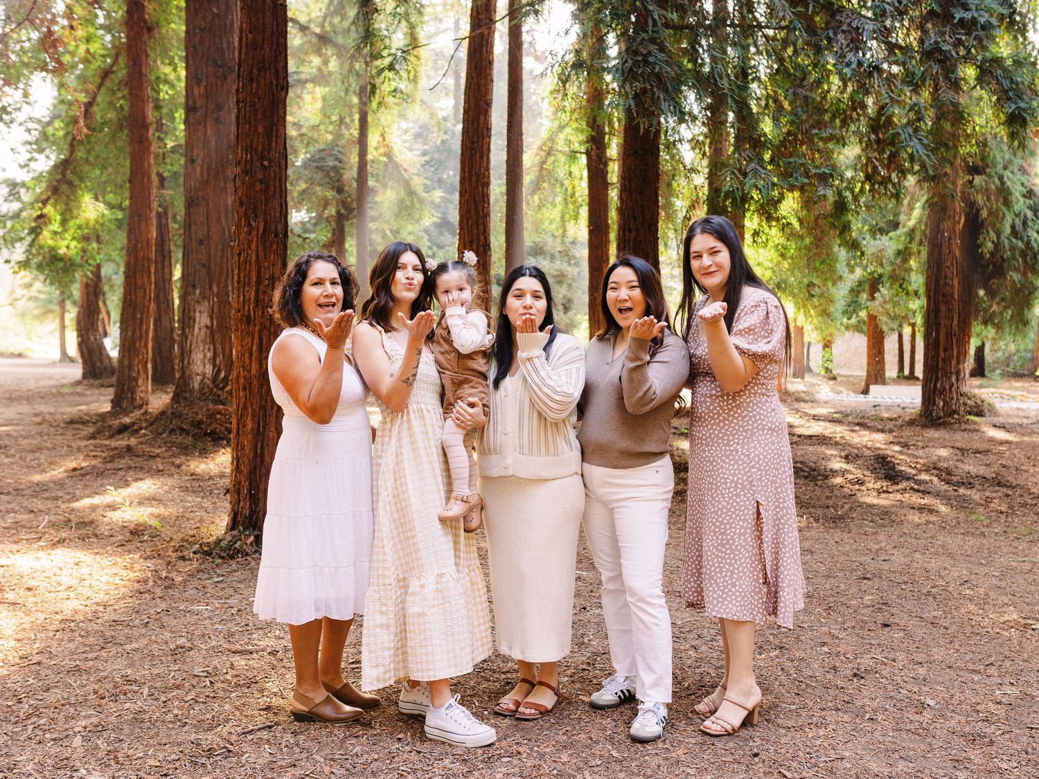 A group of friends in coordinated neutral outfits pose together on a tree-lined path.