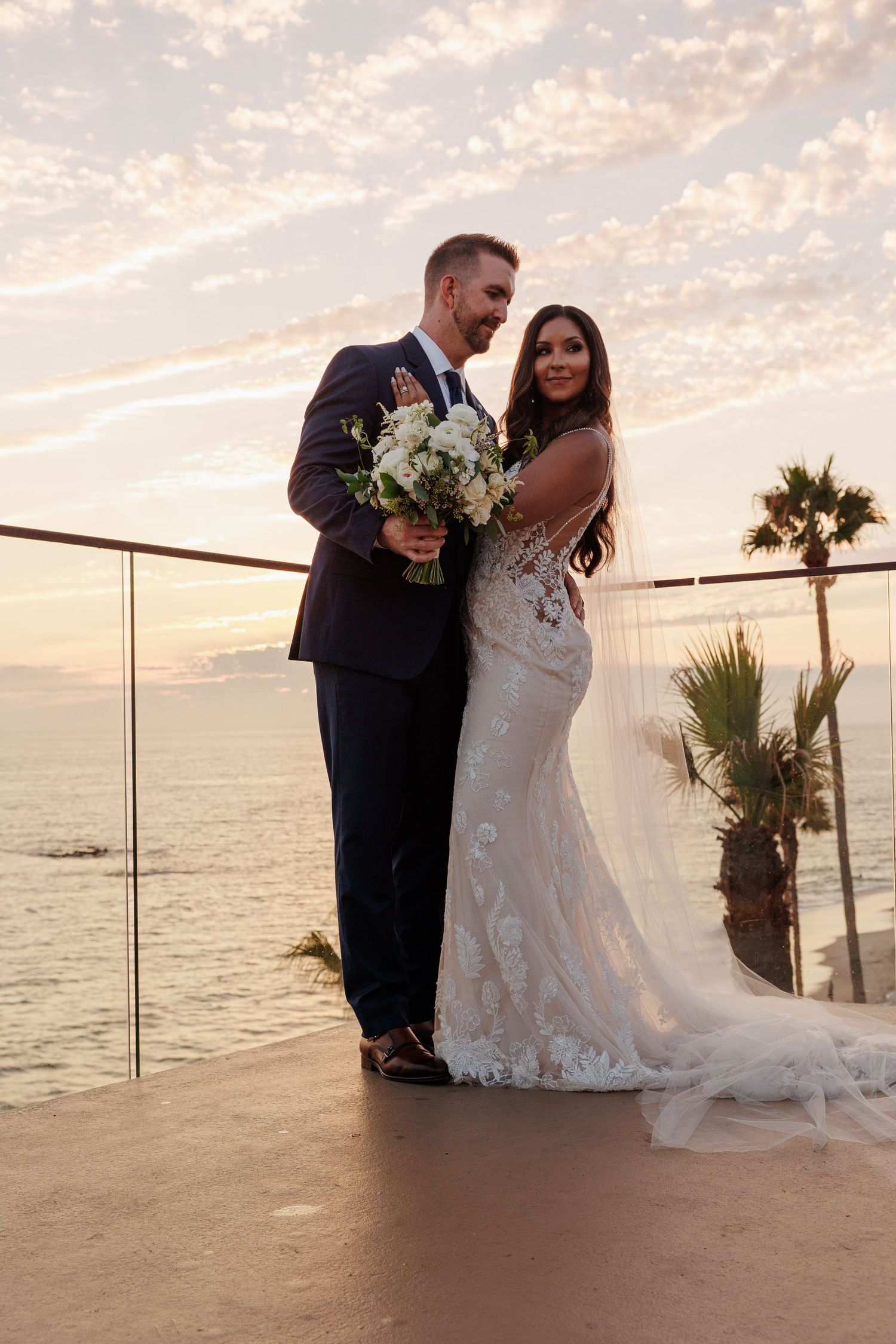 Bride and groom posing on an oceanfront balcony with palm trees during a golden sunset.