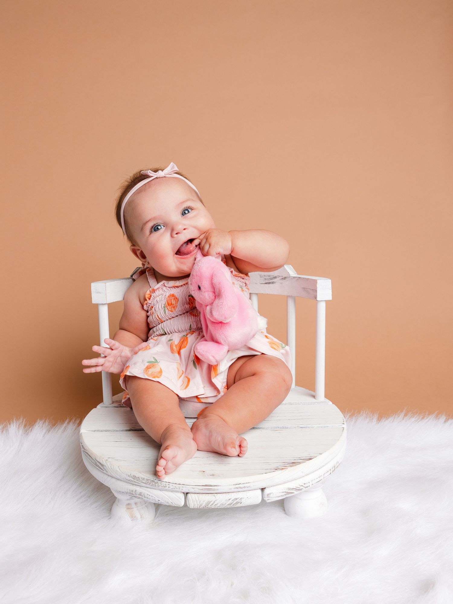 Smiling baby girl in floral romper sitting on white mini chair holding pink stuffed animal on fluffy white rug.