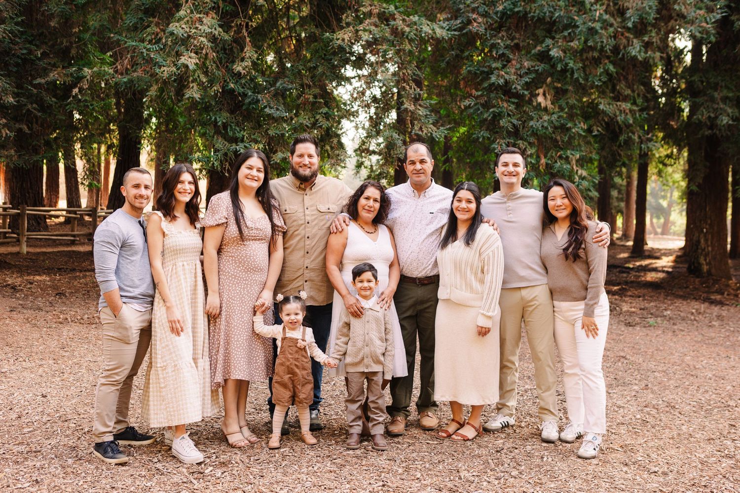 Large family group photo in neutral tones standing along wooden fence in forest setting.
