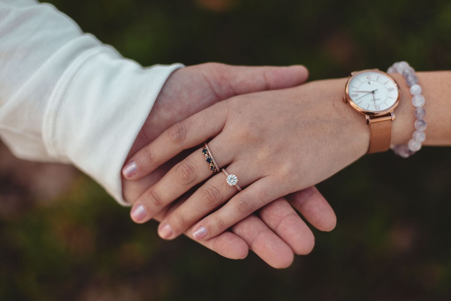 Two hands with engagement rings intertwined showing wedding bands and a leather watch.