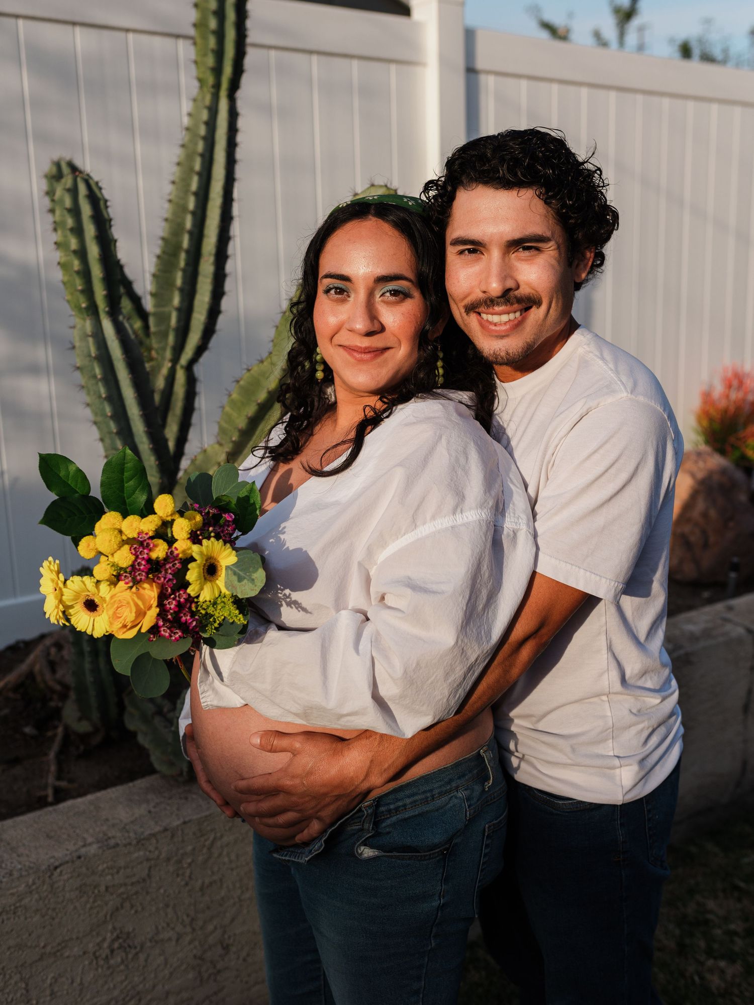 Outdoor maternity photos with yellow sunflowers against white fence and cacti.