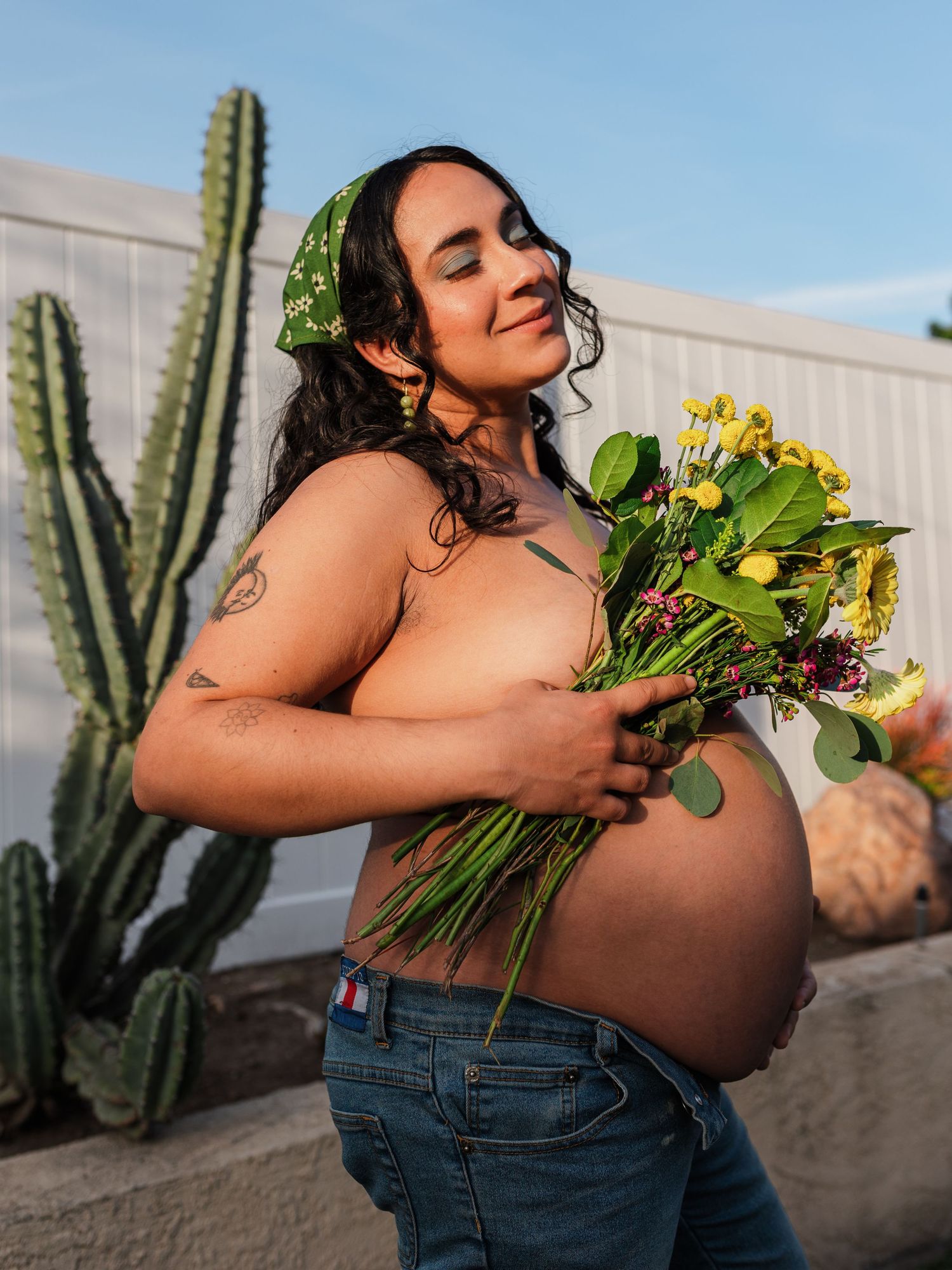 A pregnant person holding yellow flowers poses next to a tall cactus outdoors.