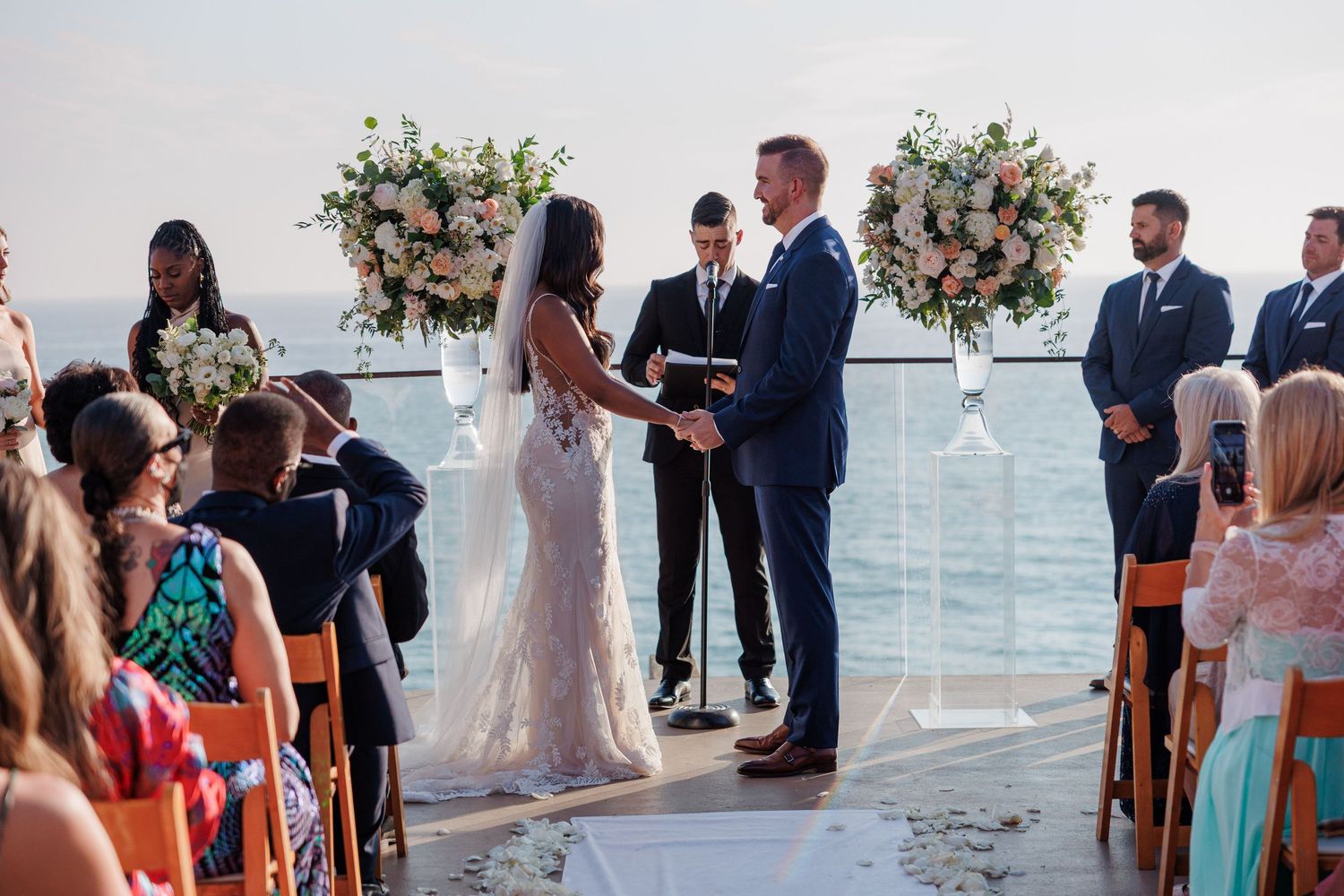 Couple exchanging vows at outdoor ocean-view wedding ceremony with floral arrangements and seated guests.
