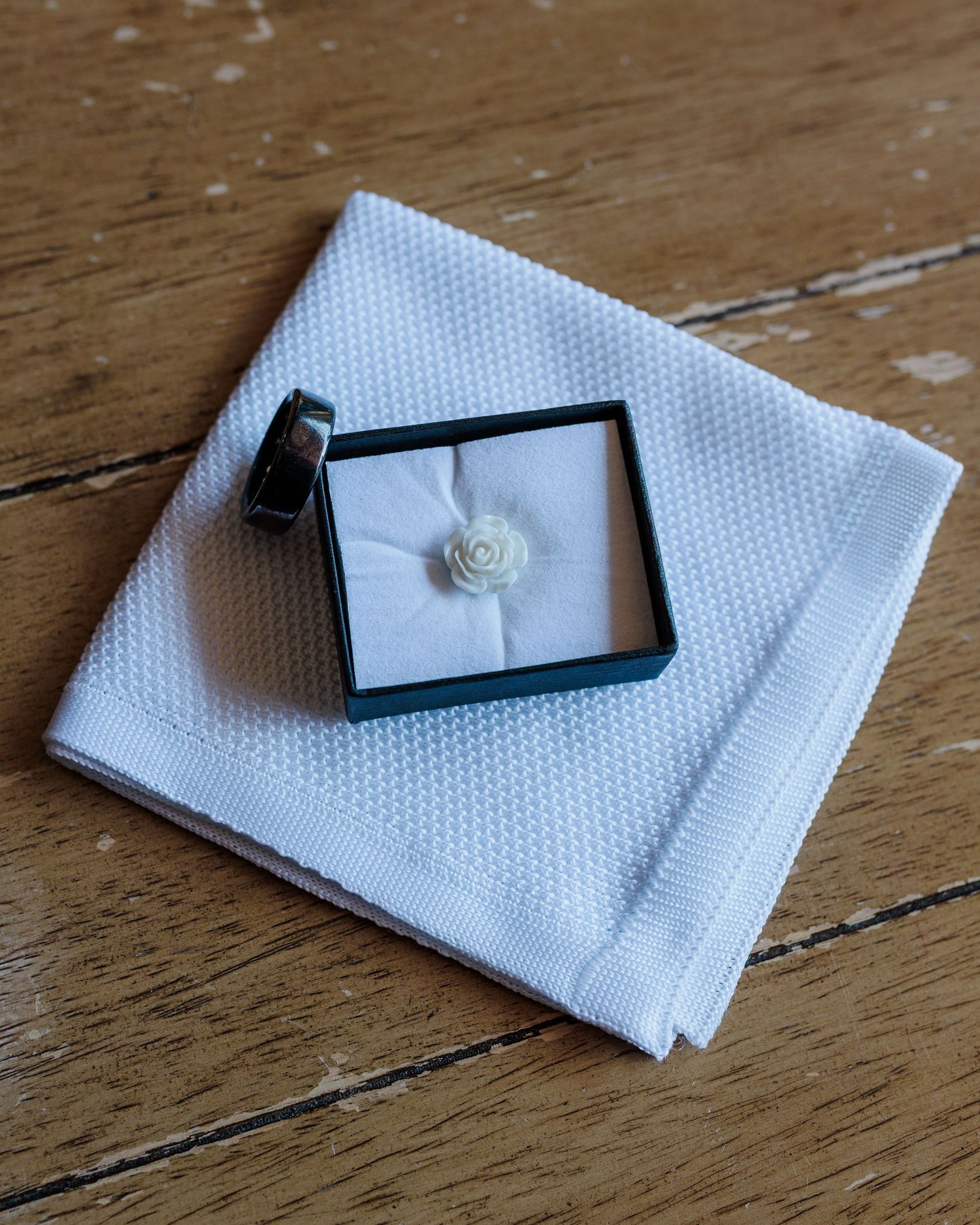 White pocket square, black ring, and white rose lapel pin in teal box on wooden surface for wedding day accessories.