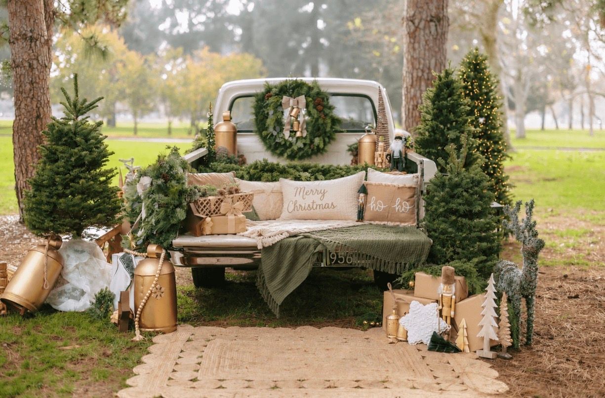 Vintage truck bed decorated with Christmas pillows, wreath, and surrounded by small pine trees and holiday gifts outdoors.