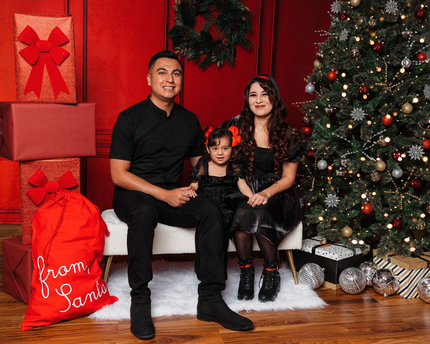 Family of three posing for a Christmas photo with a decorated tree, red gift boxes, and a Santa sack.