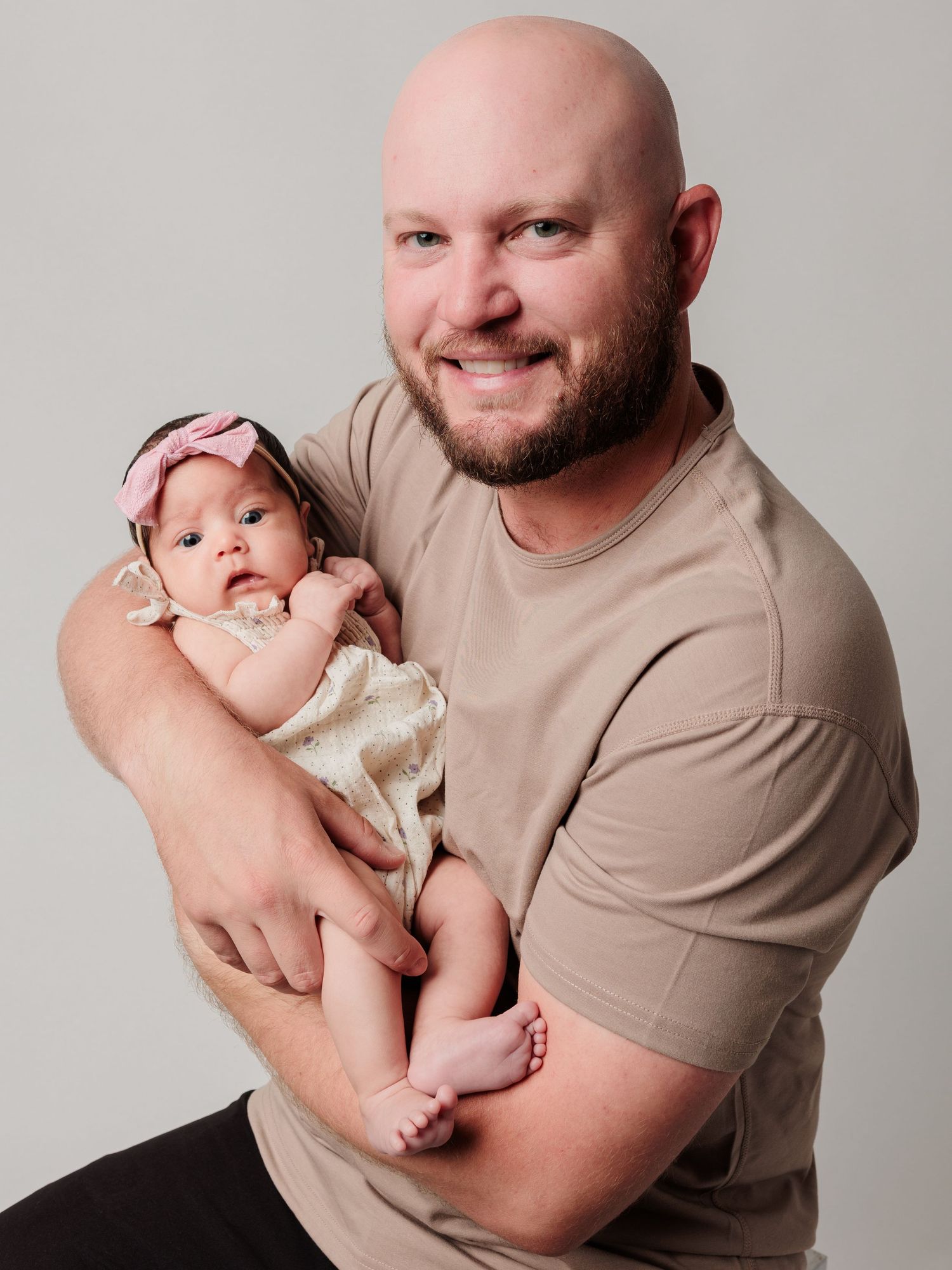 A parent in a tan shirt holds a small baby wearing a pink bow headband in a studio portrait.