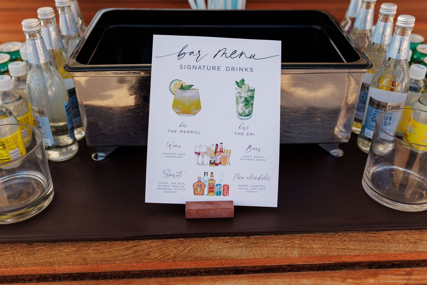 Bar menu sign displaying signature drinks with illustrated cocktails, surrounded by bottled beverages on a wooden table.