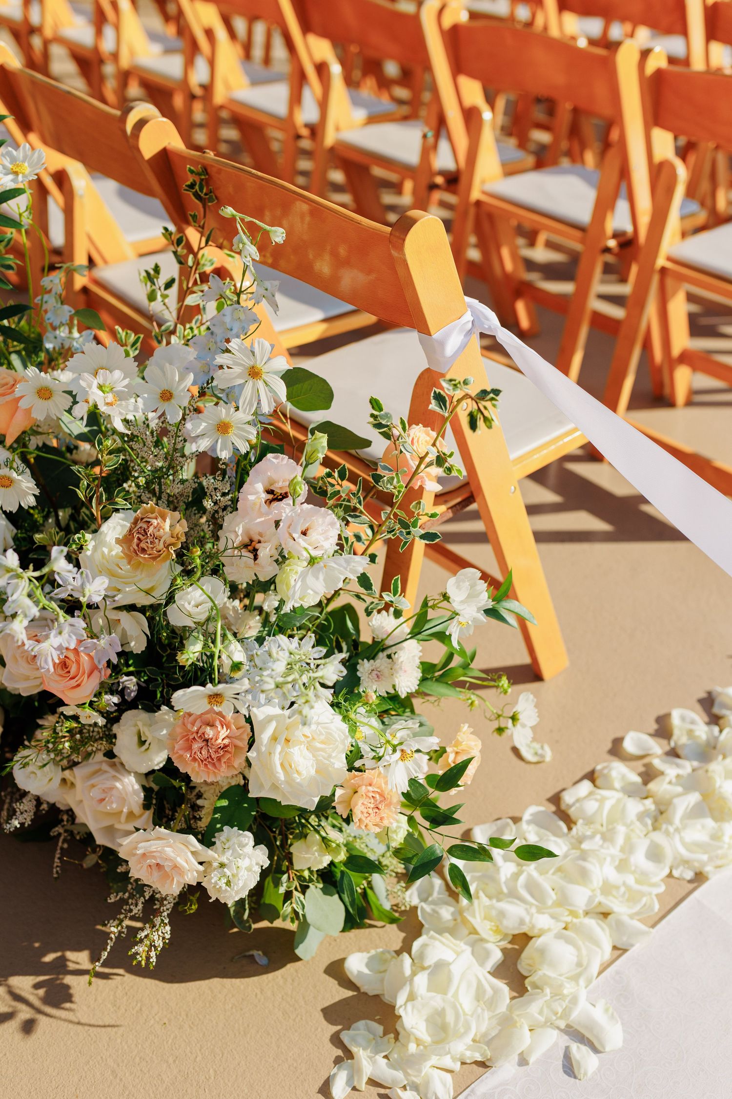 Elegant outdoor wedding ceremony with orange chairs, white rose petals, and lush floral arrangements in blush and white tones.