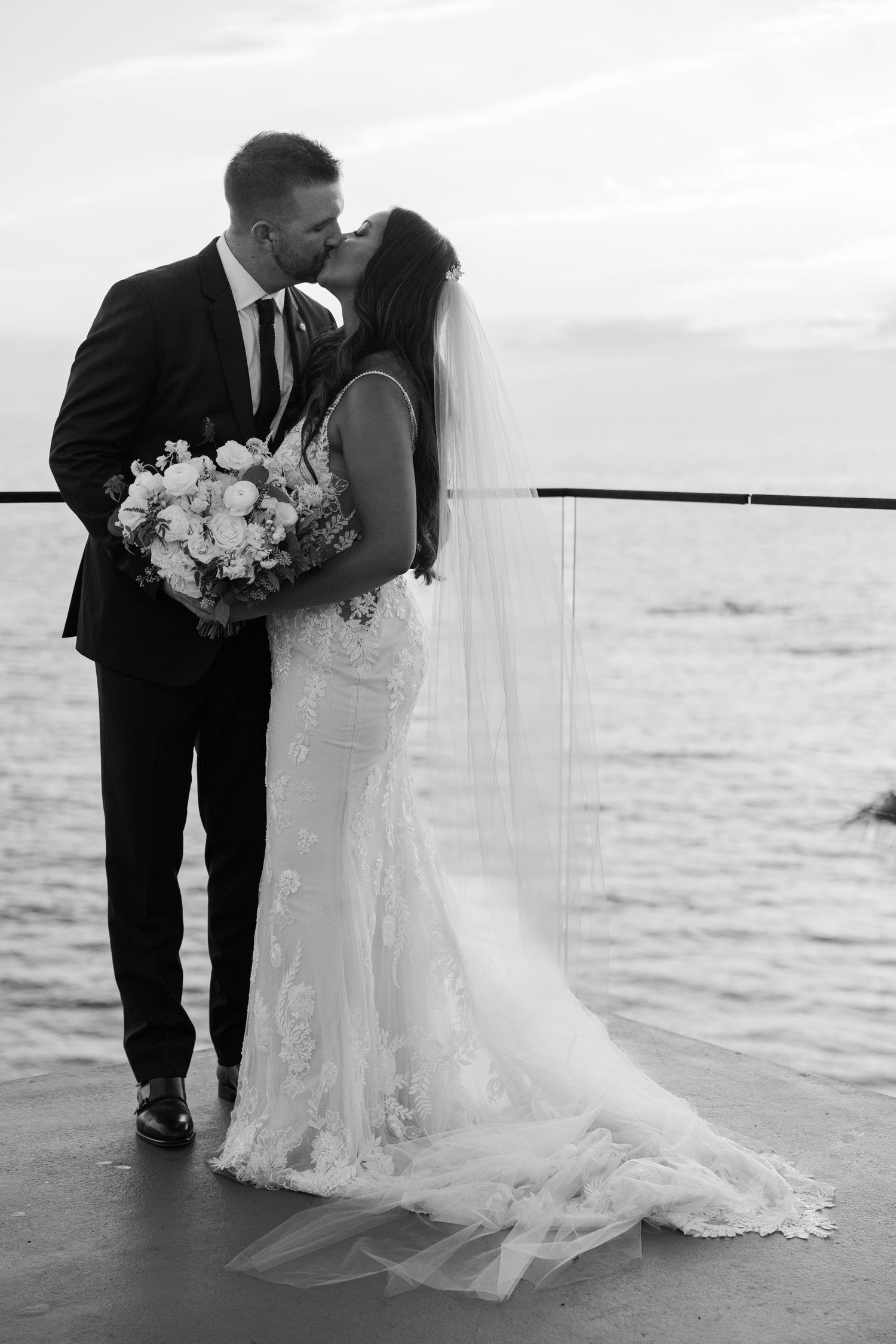 Black and white photo of bride and groom sharing a kiss on a waterfront terrace at sunset.