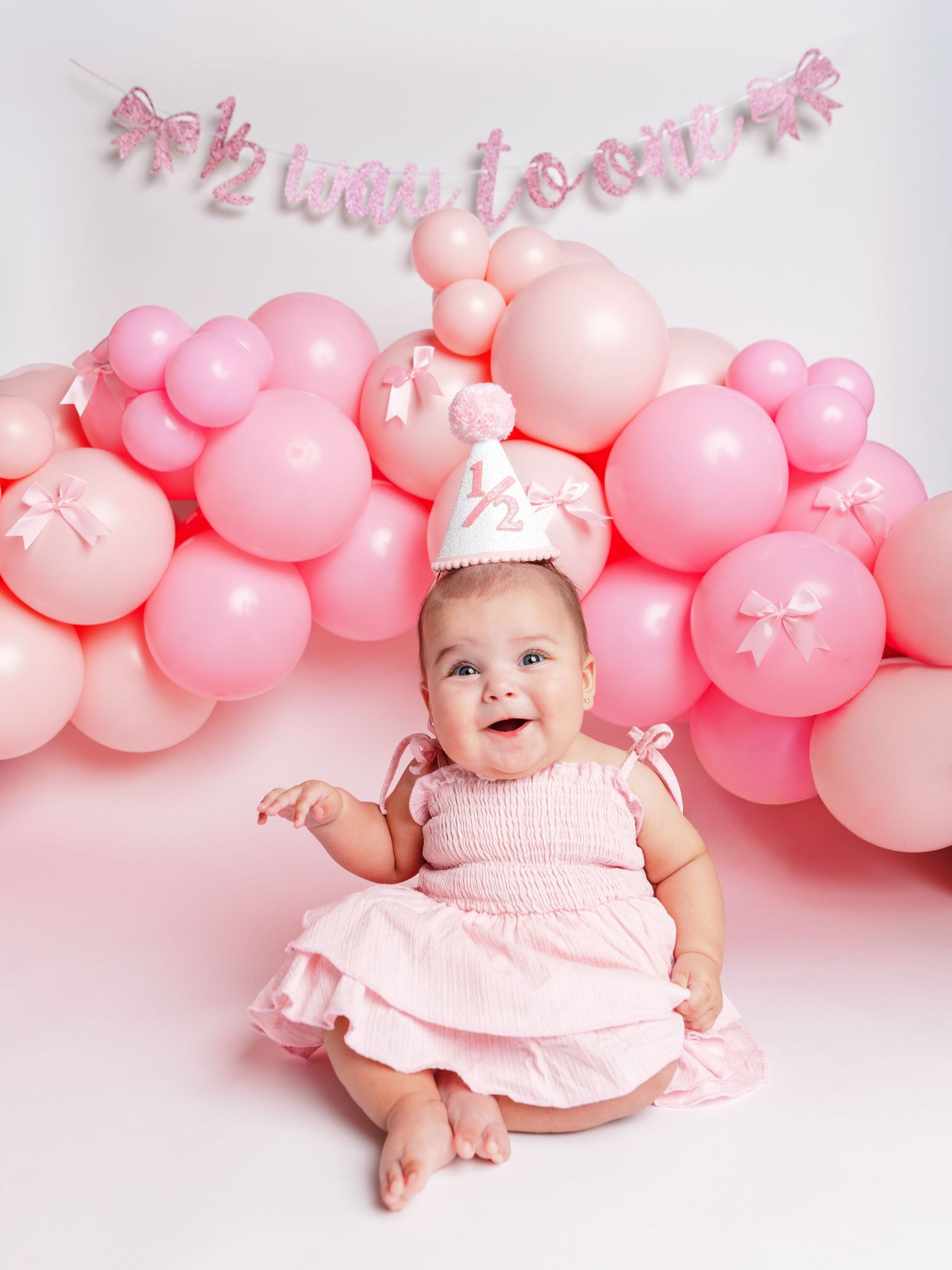 A smiling baby in a pink dress and half birthday crown sits before a pink balloon arch and banner.