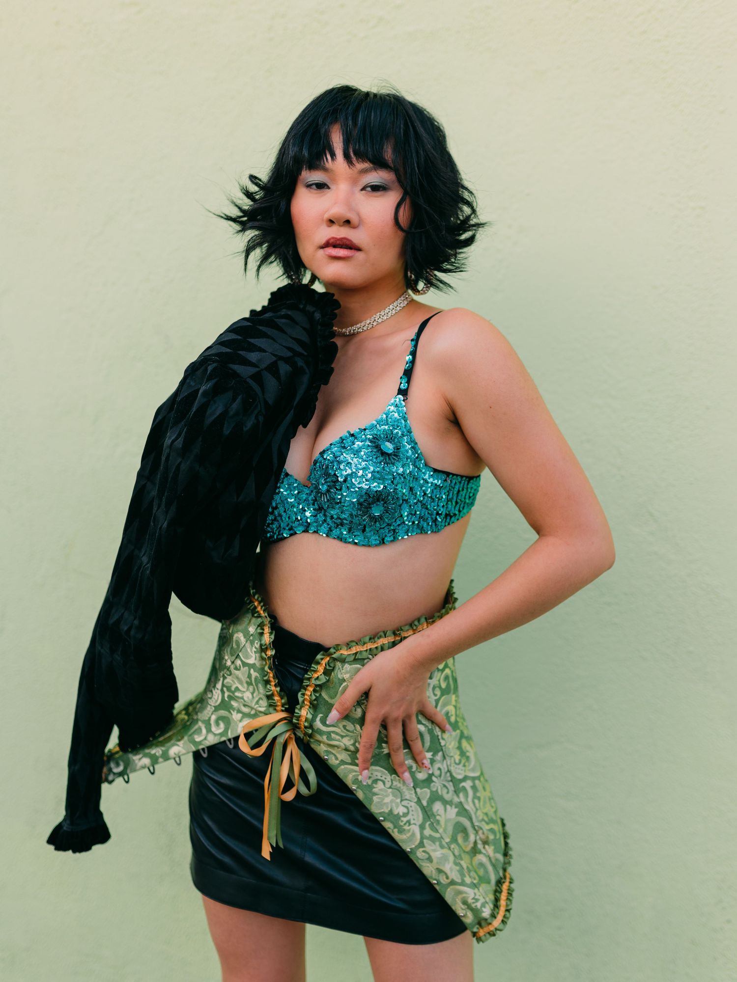 Woman in teal sequin bralette, green brocade skirt, and black fur jacket poses against a light green wall.