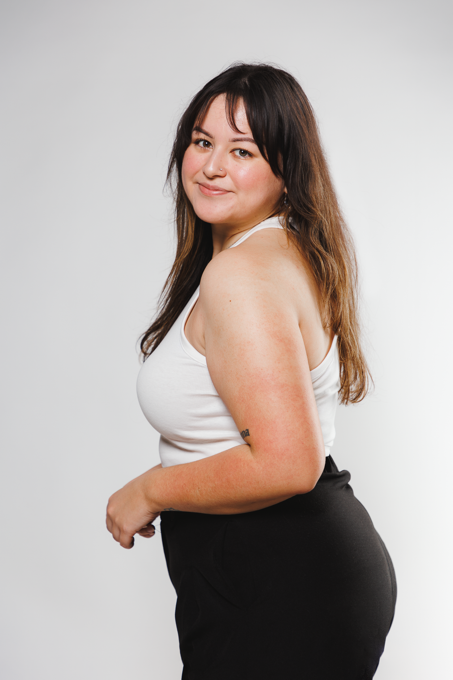 A confident woman in a white tank top and black pants poses against a light gray background.