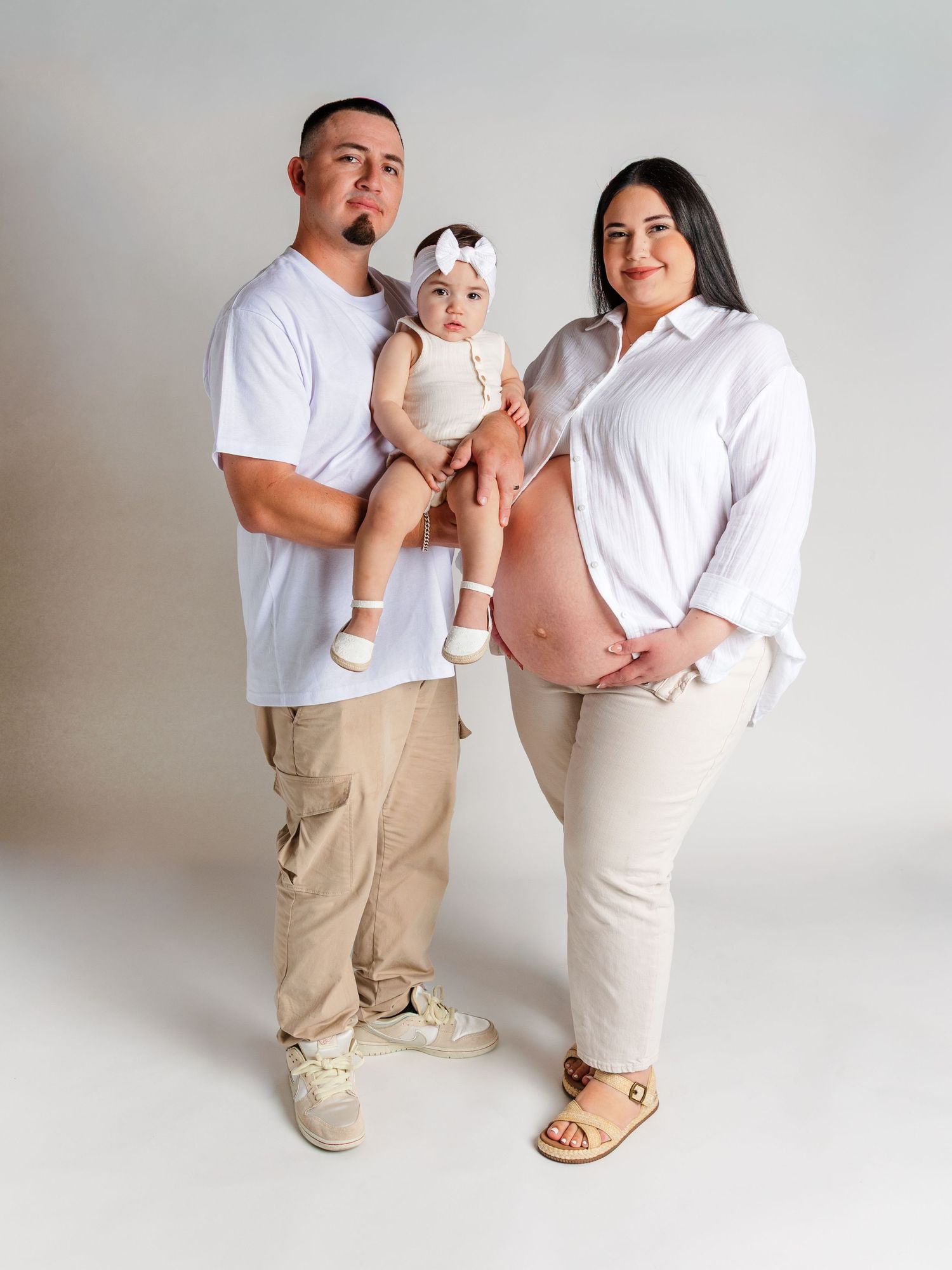 A couple in matching white shirts and beige pants pose with their baby girl in a bright studio maternity photo.