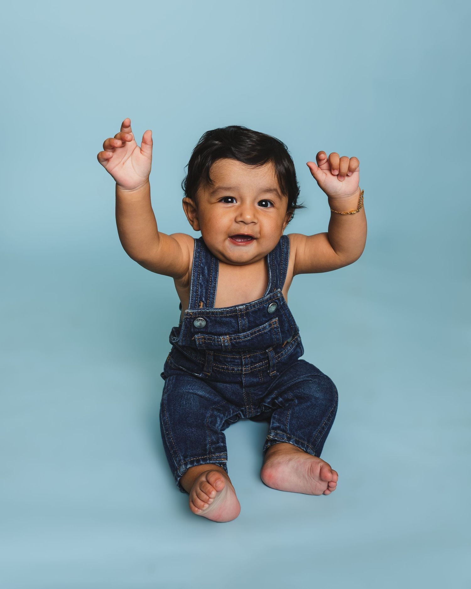 Happy baby in denim overalls raising arms joyfully against light blue background.