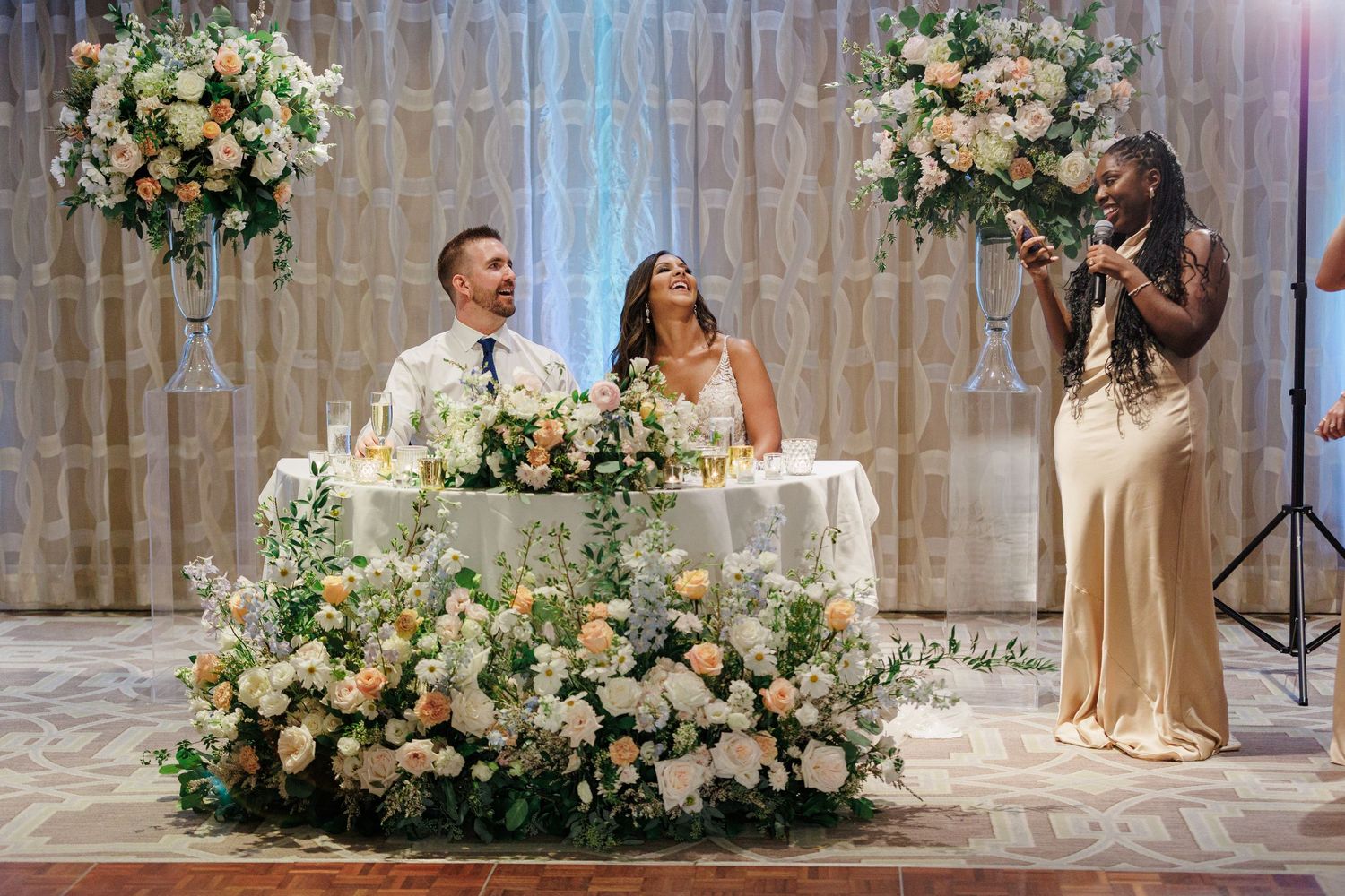 Bride and groom laugh at sweetheart table adorned with lush floral arrangements during reception.