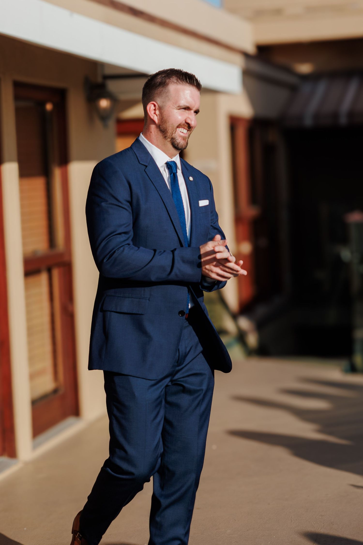 Smiling groom in navy blue suit clapping hands outdoors on sunny wedding day.