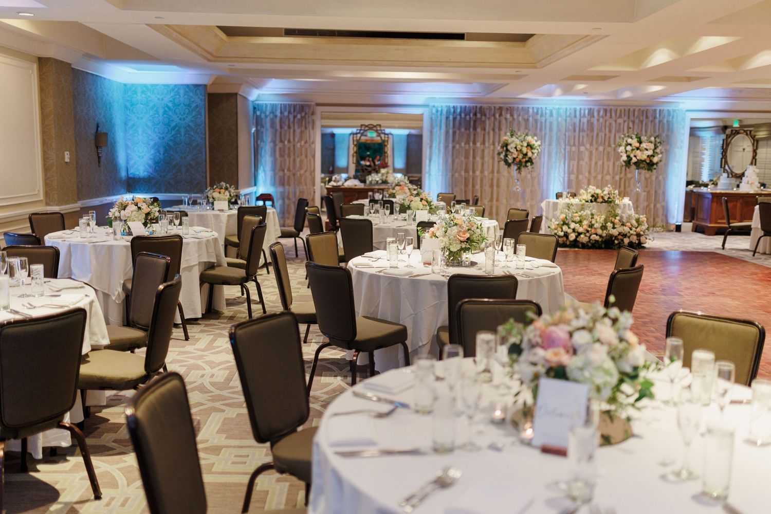 Elegant wedding reception ballroom with round tables, white floral centerpieces, and soft blue uplighting.
