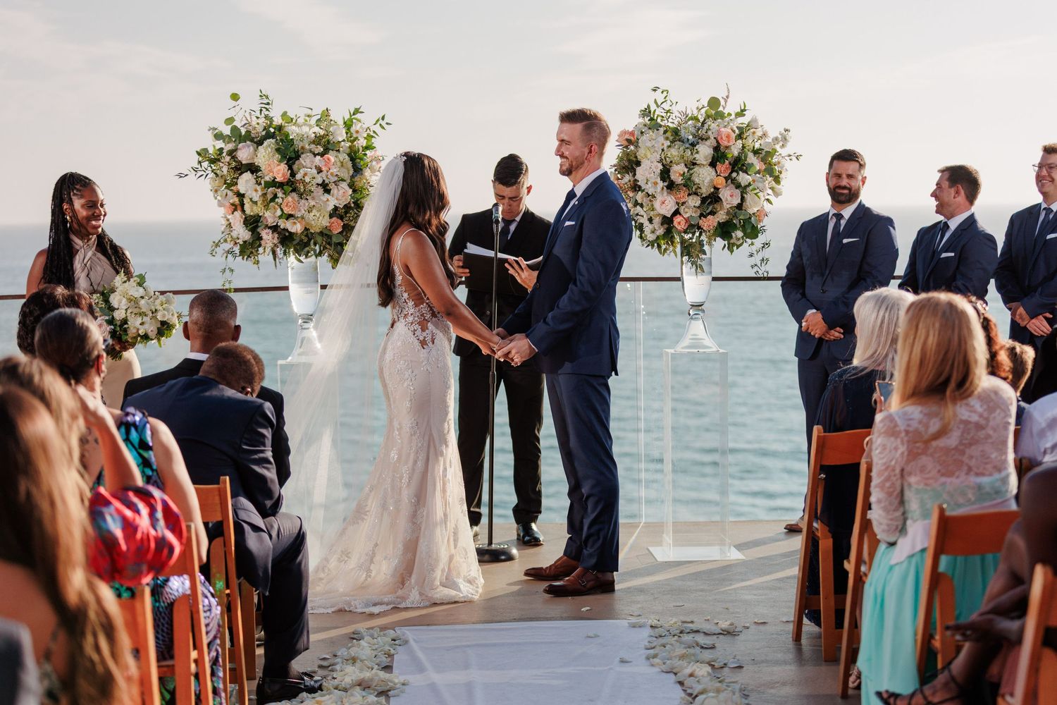 Couple holding hands at altar during outdoor waterfront wedding ceremony with guests and floral decor.