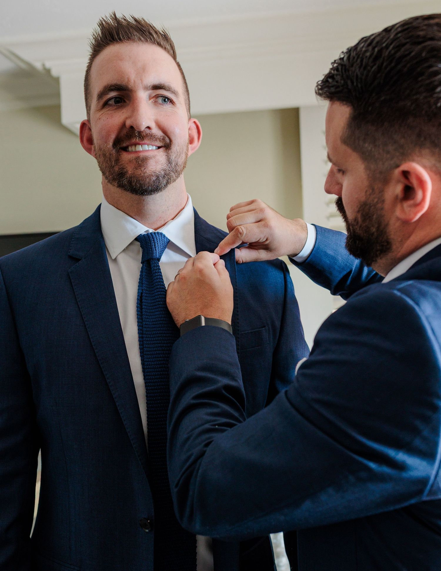 Groomsman helping groom with boutonniere on navy suit lapel while smiling during wedding prep.