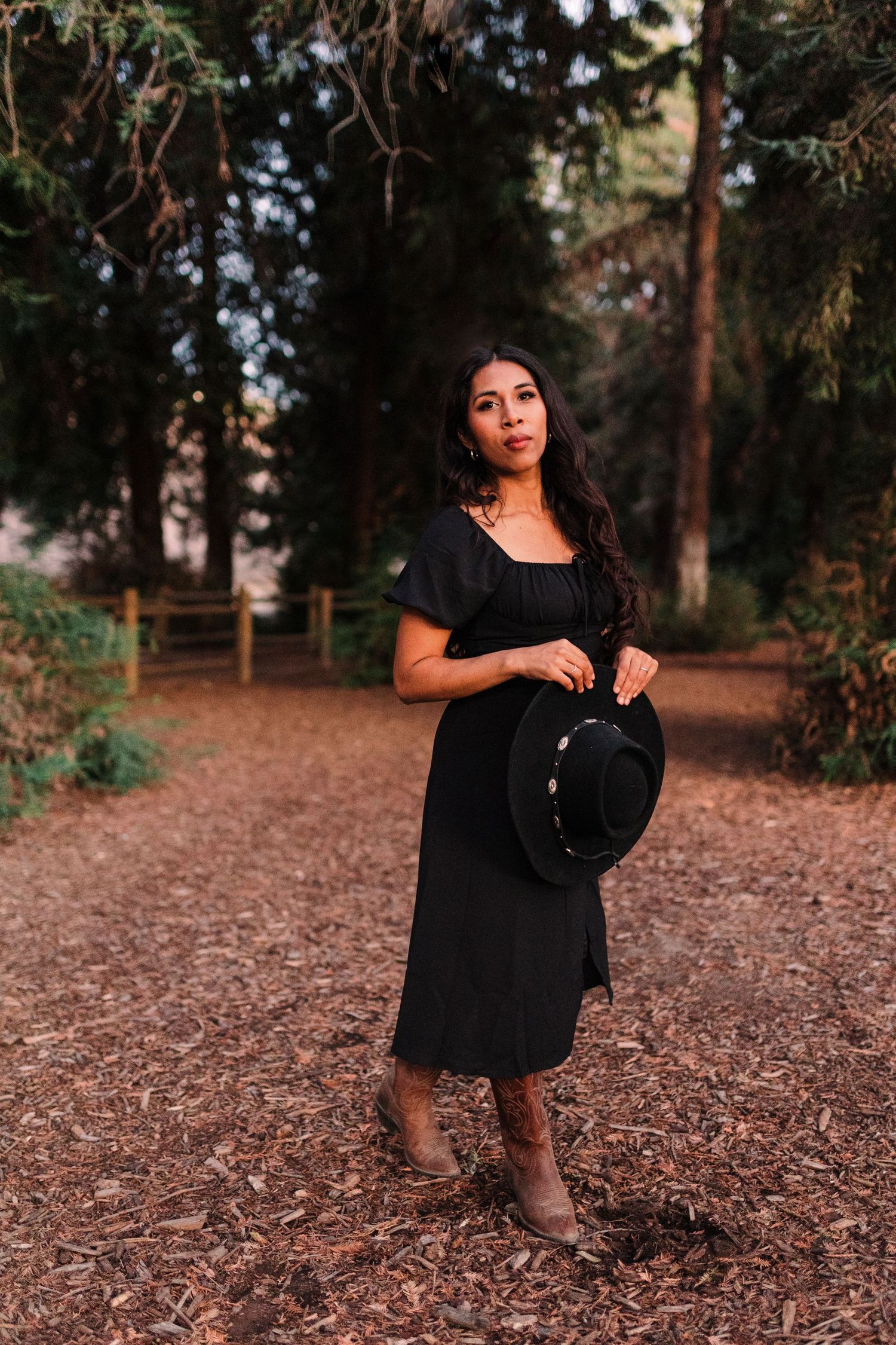 Woman in black dress holding cowboy hat standing on forest path among tall trees.