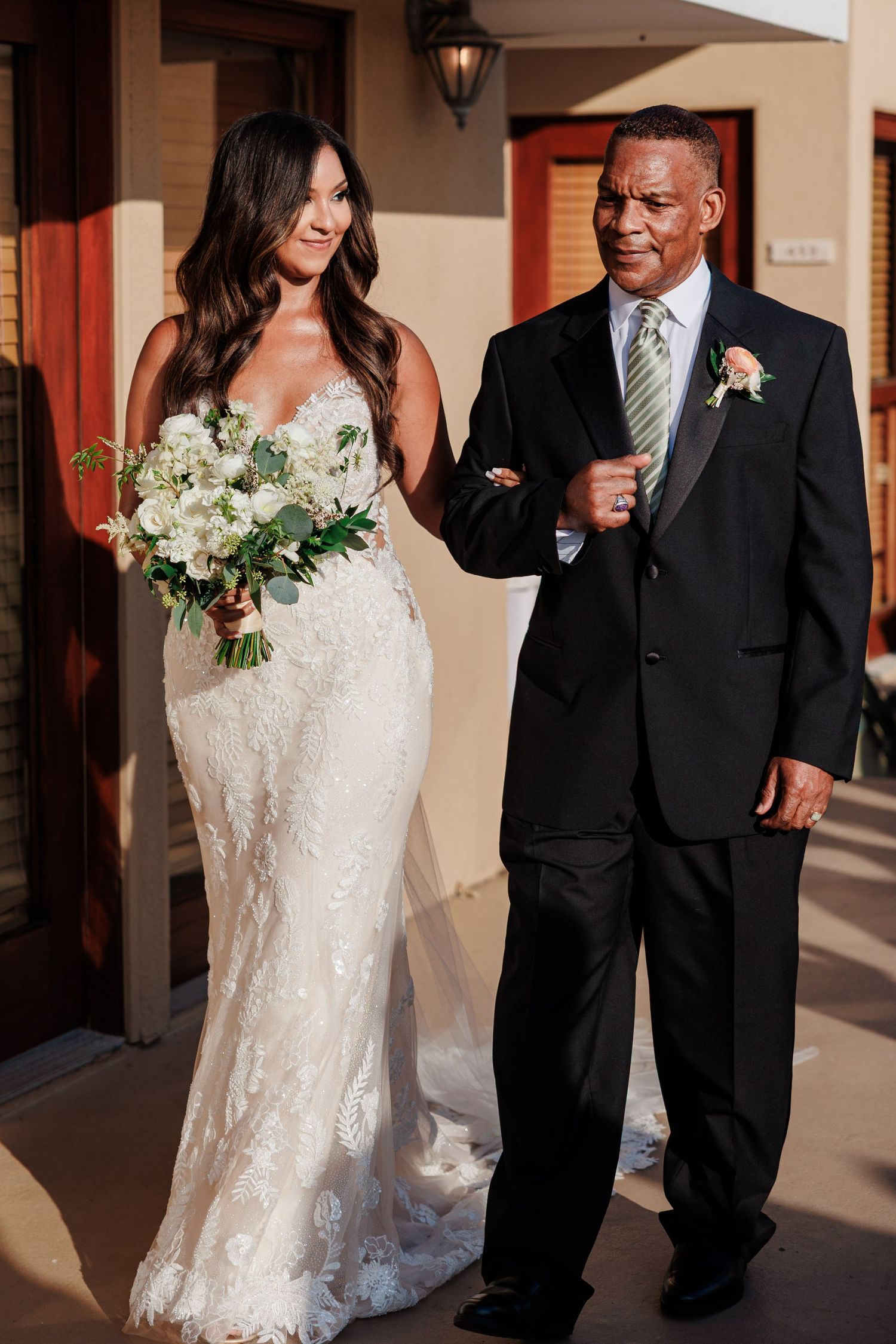 Bride in lace mermaid gown holding white bouquet walks with Father in black suit.
