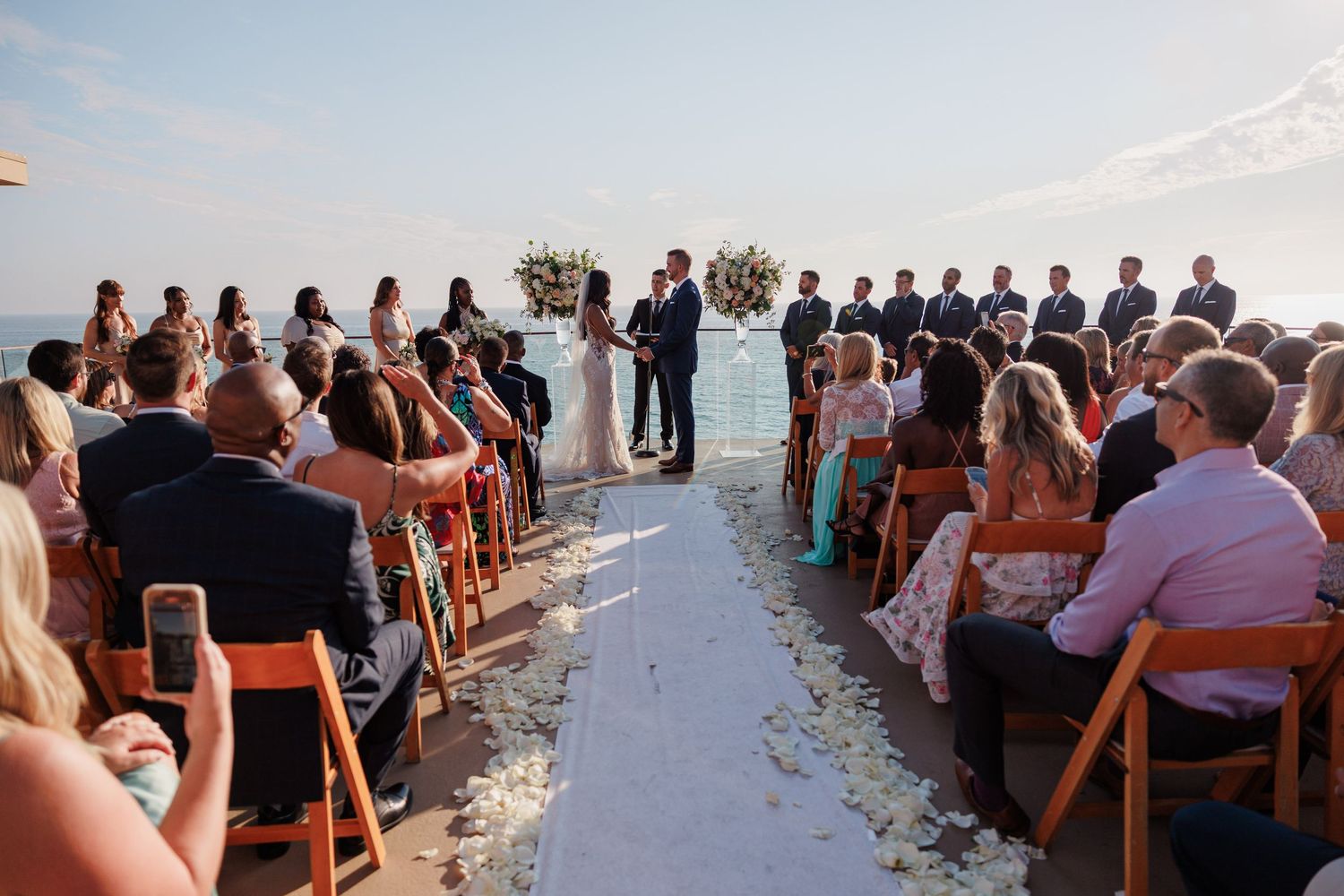 Outdoor beach wedding ceremony with guests seated facing couple at floral altar overlooking ocean.