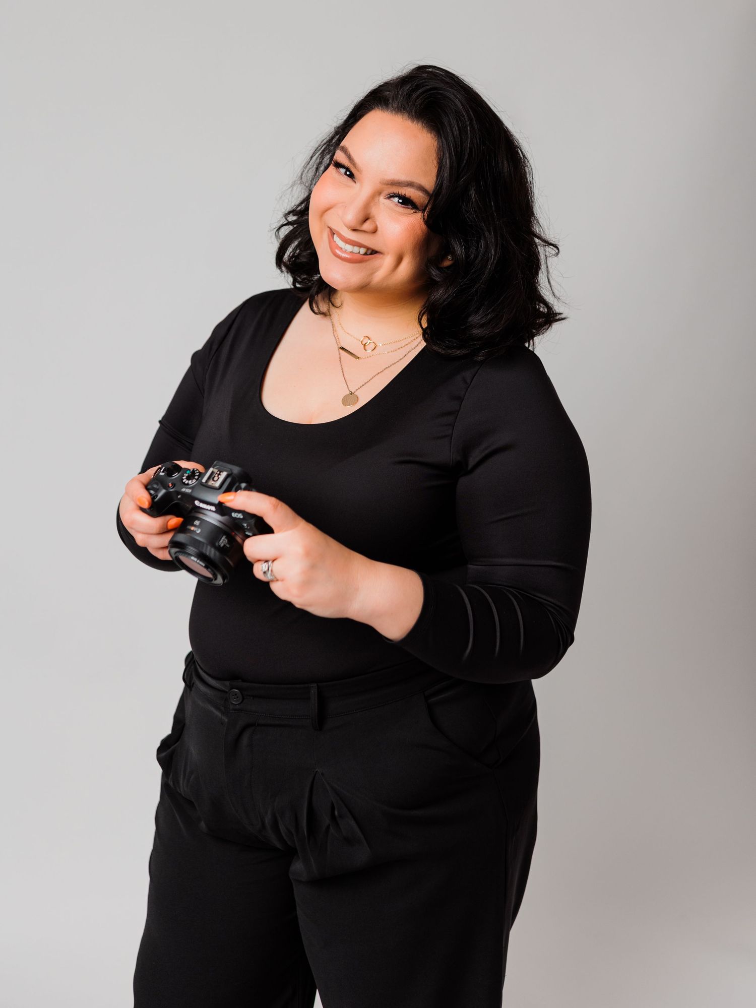Studio portraits in all-black outfit holding camera against light gray background.