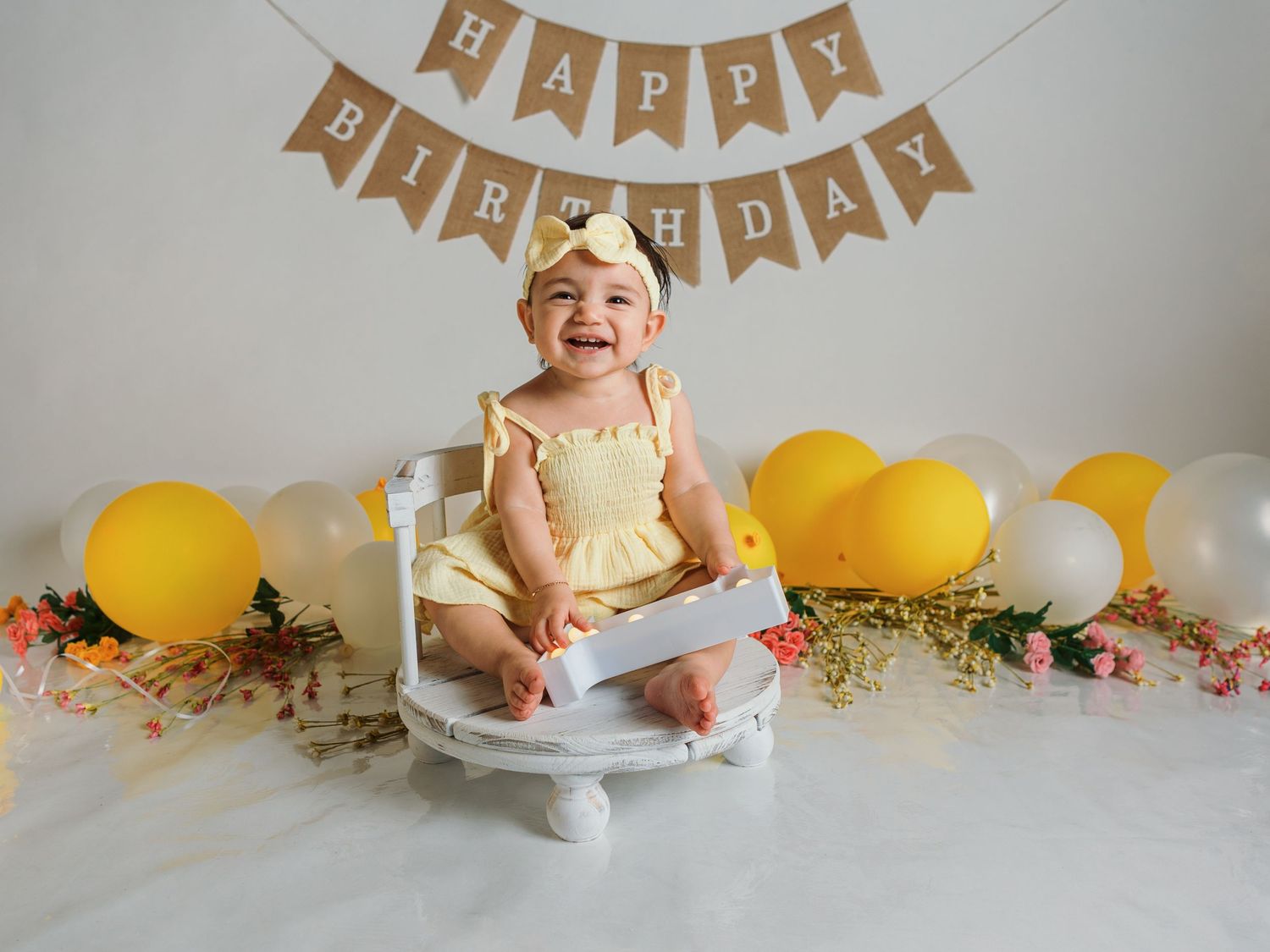 First birthday celebration setup with kraft paper banner, yellow balloons, and white cake on pedestal.