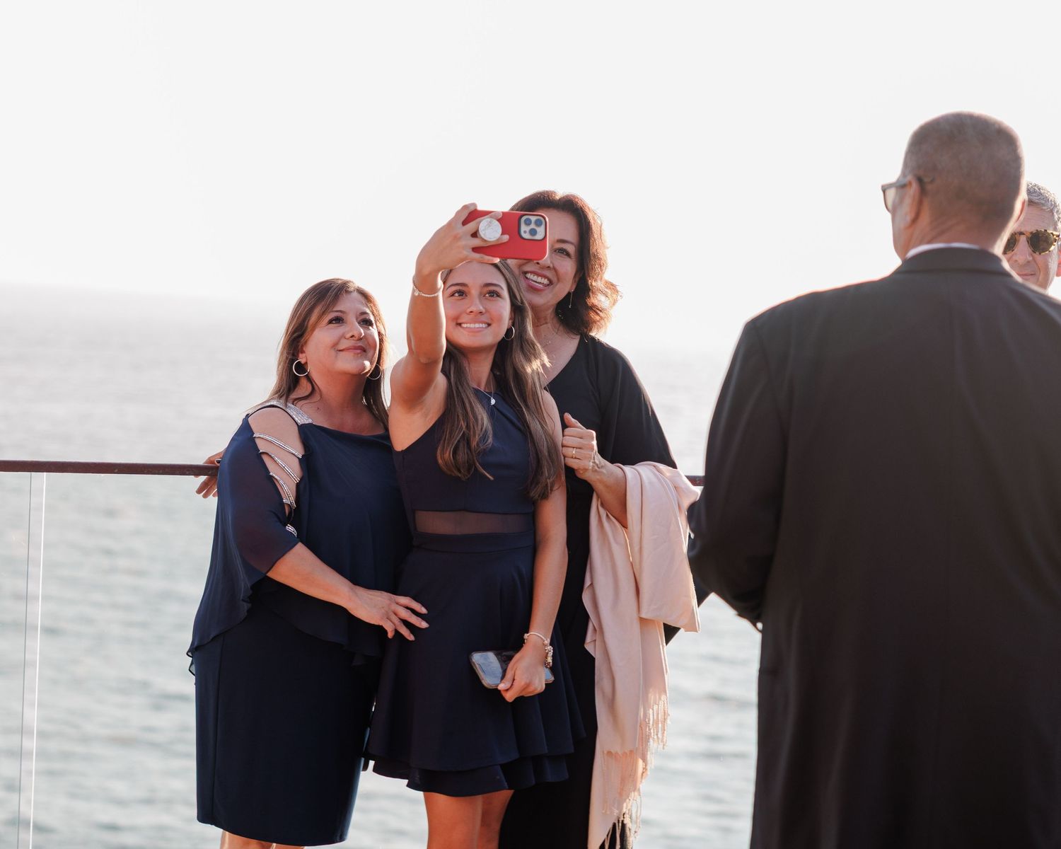 Three women take a selfie with a red camera on a cruise ship deck overlooking the ocean at sunset.
