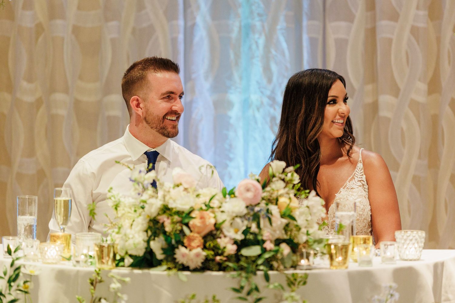 Bride and groom smiling at sweetheart table adorned with lush white and blush floral arrangements at elegant wedding reception.