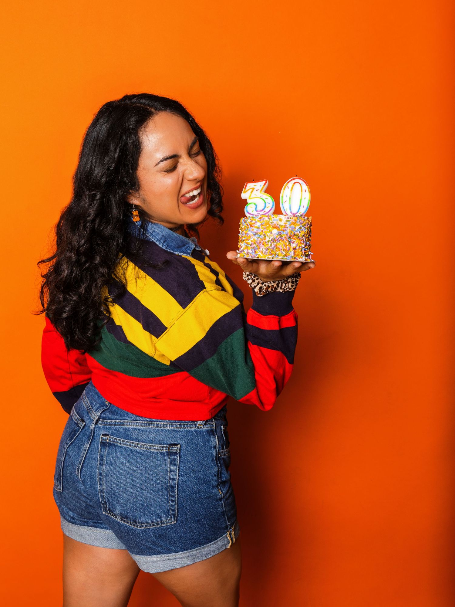 Person wearing colorful retro-inspired outfit with denim shorts and color block sweater poses against vibrant orange background.
