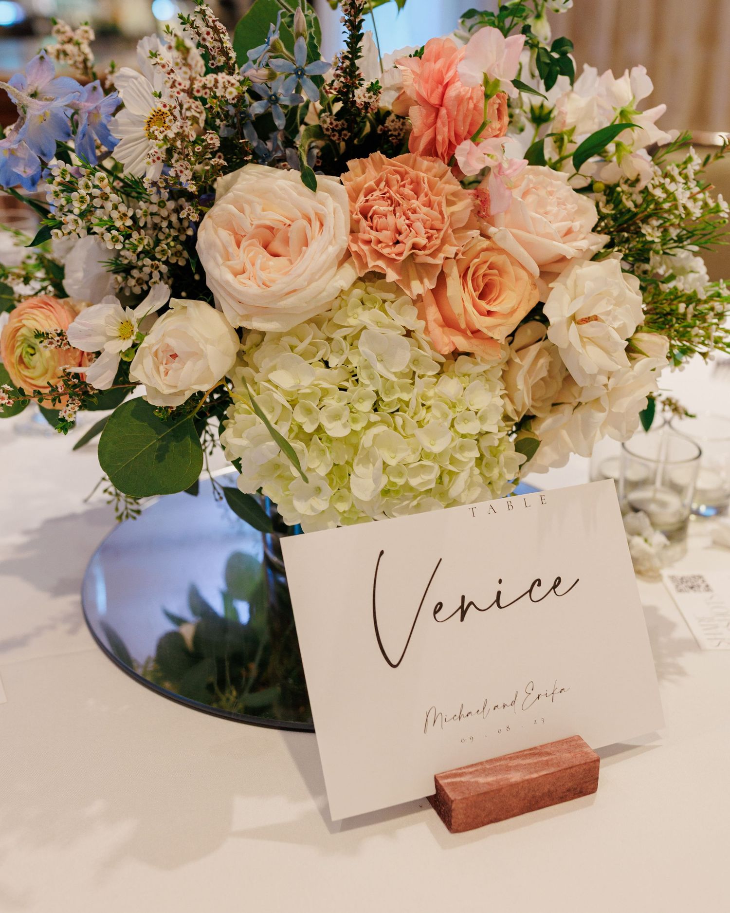 Elegant wedding table centerpiece with peach roses and hydrangeas beside a Venice table name card in wooden holder.