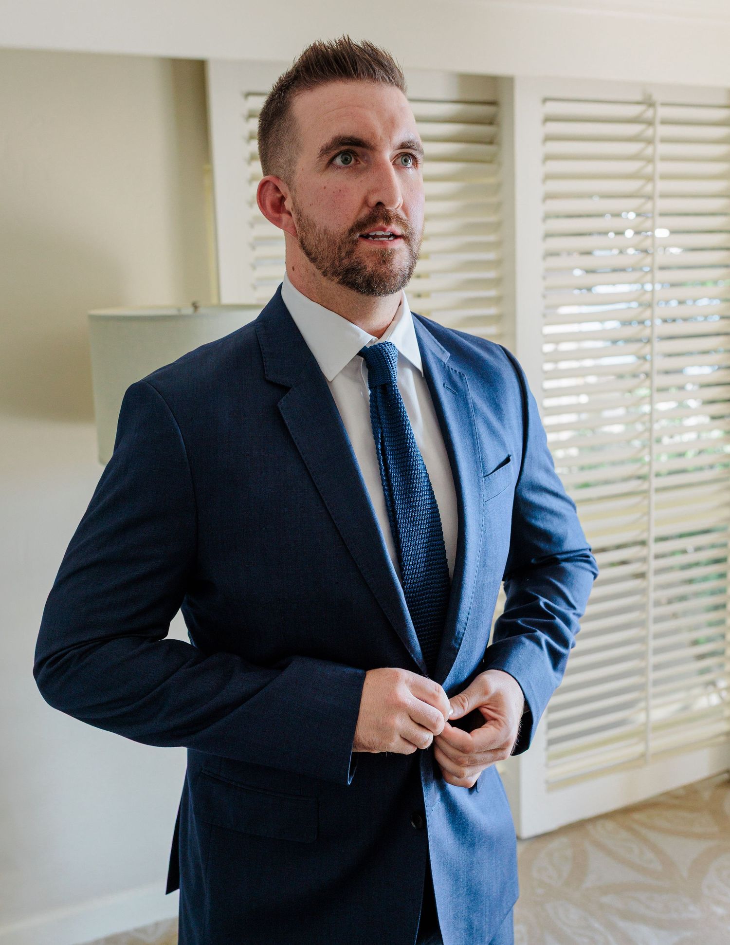 Groom in navy blue suit and tie buttoning jacket while getting ready for wedding ceremony.