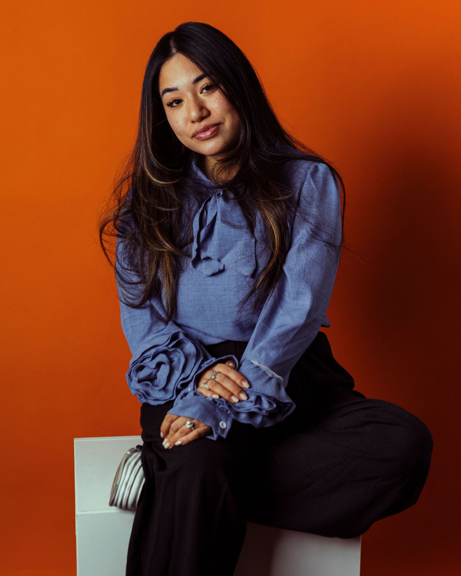 Woman in blue ruffled blouse and black pants sits confidently against vibrant orange background.