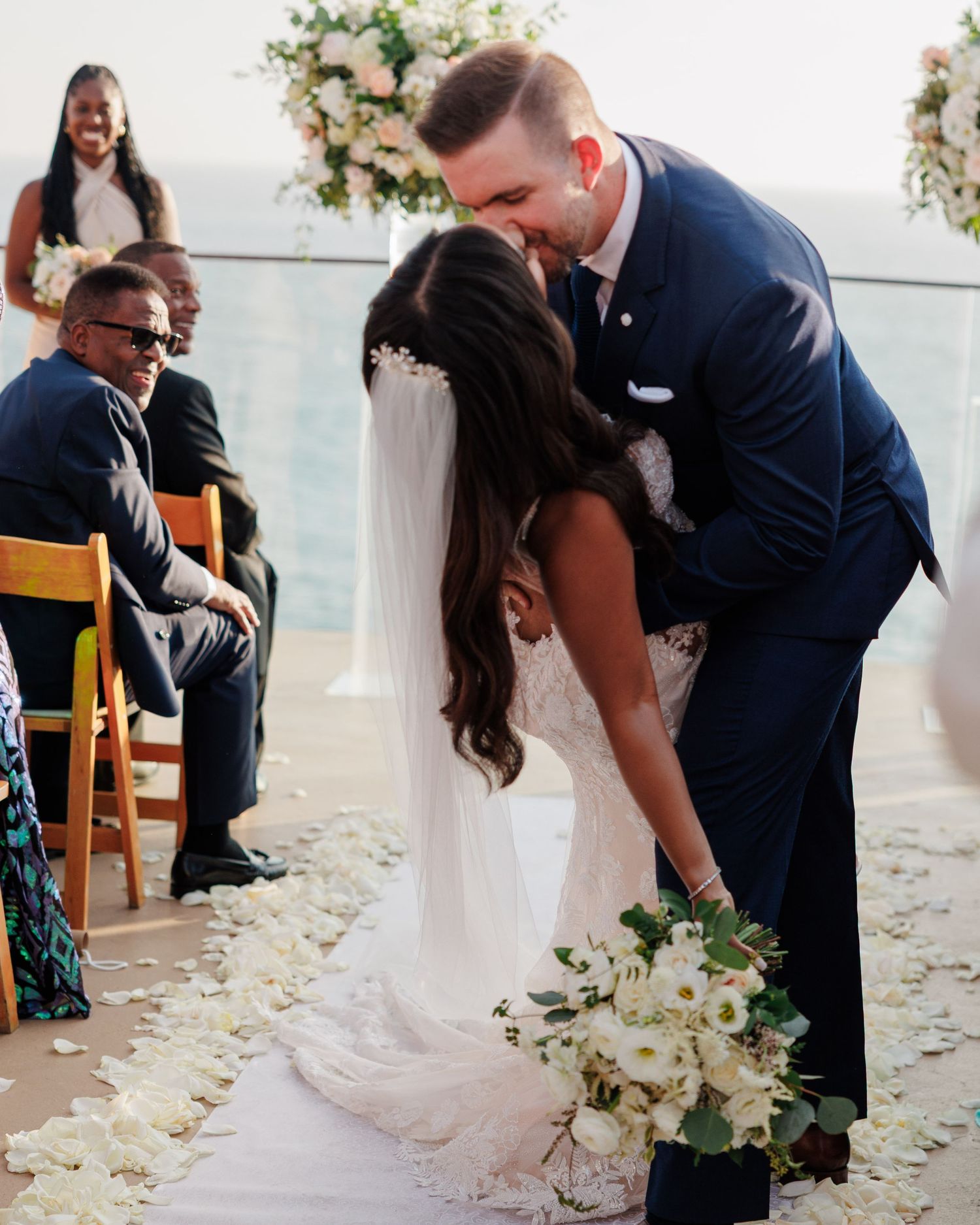 Groom in navy suit leaning down to kiss bride in lace gown at outdoor wedding ceremony.