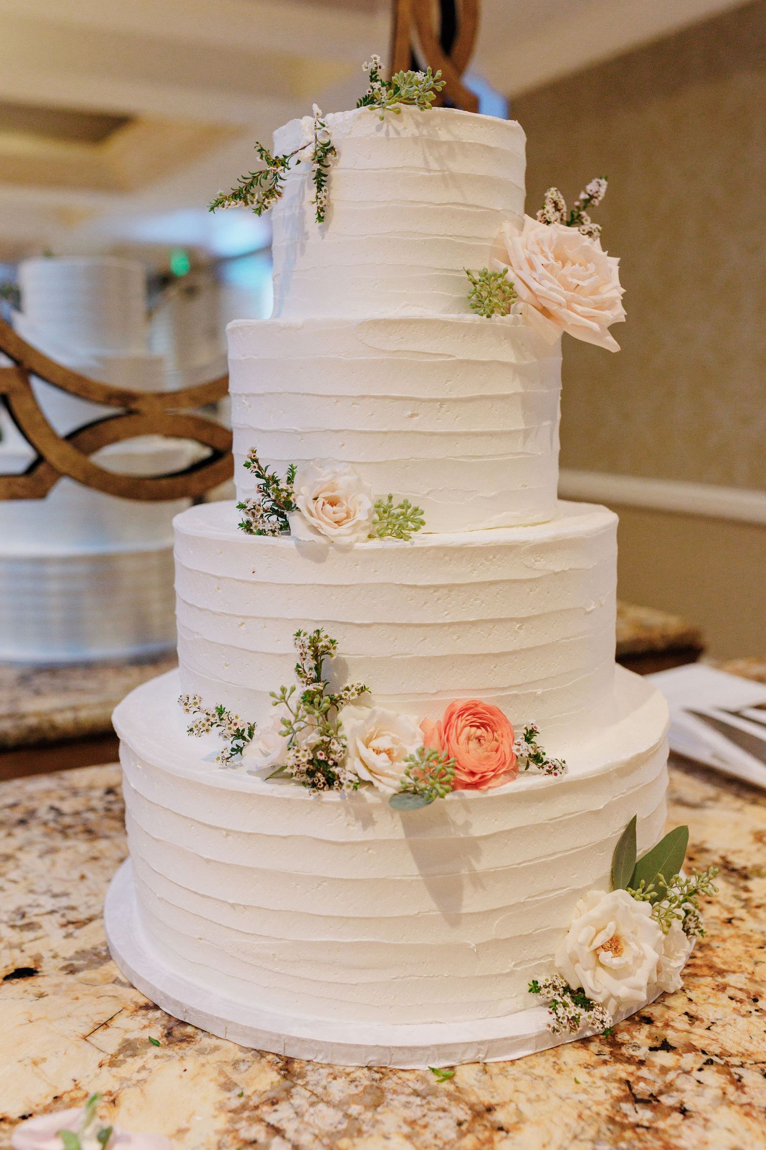 Four-tier white wedding cake with textured frosting and pink and white floral decorations.