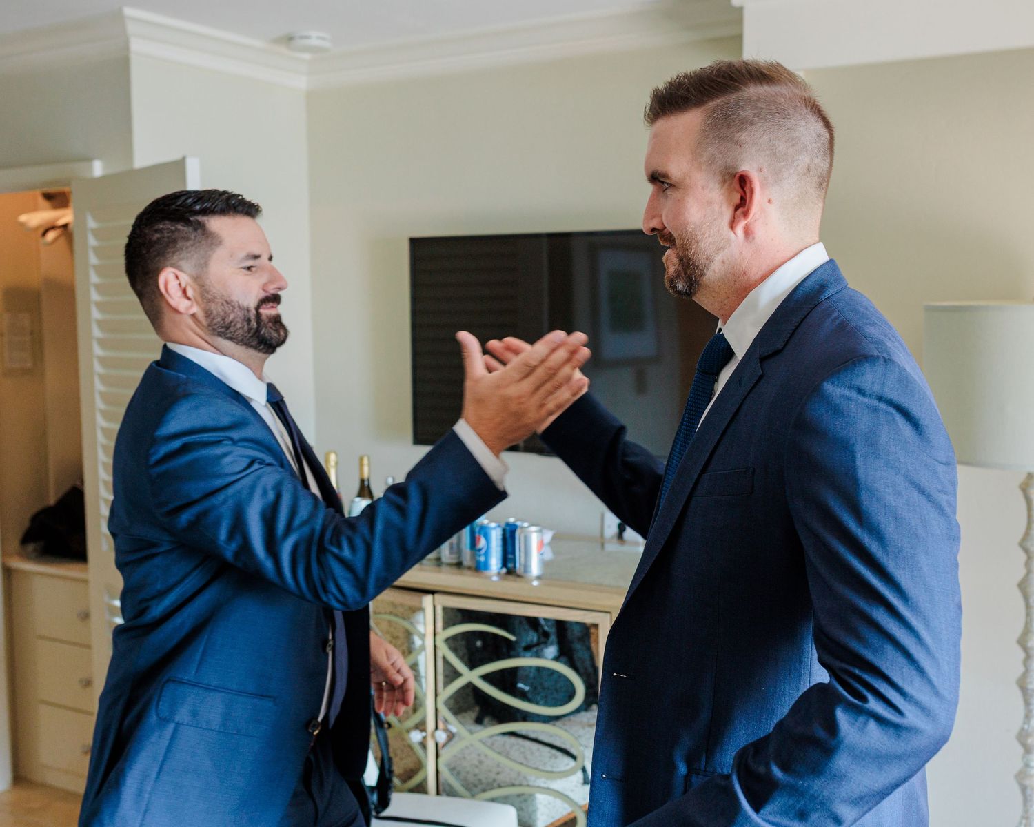 Two men in navy blue suits share a joyful handshake moment, likely at a wedding celebration indoors.