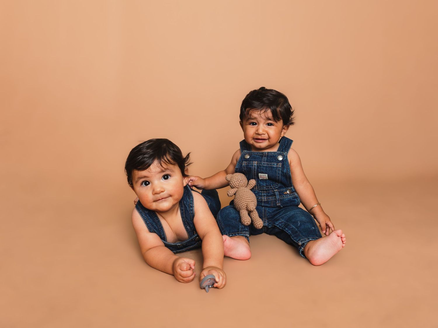 Two toddlers in denim overalls sitting together with a plush toy on peach background.