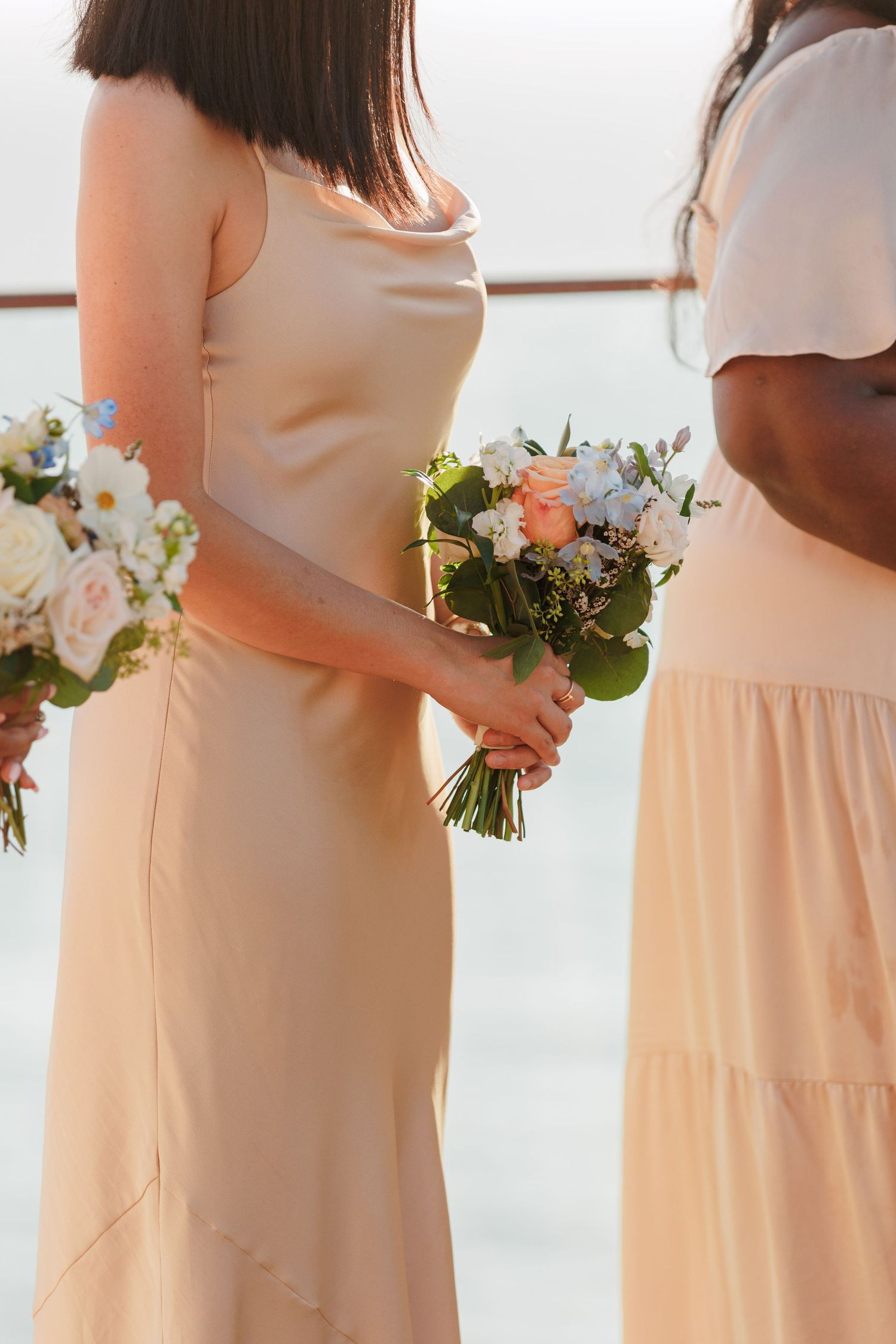 Bridesmaids in champagne satin dresses hold white and peach floral bouquets at outdoor wedding ceremony.