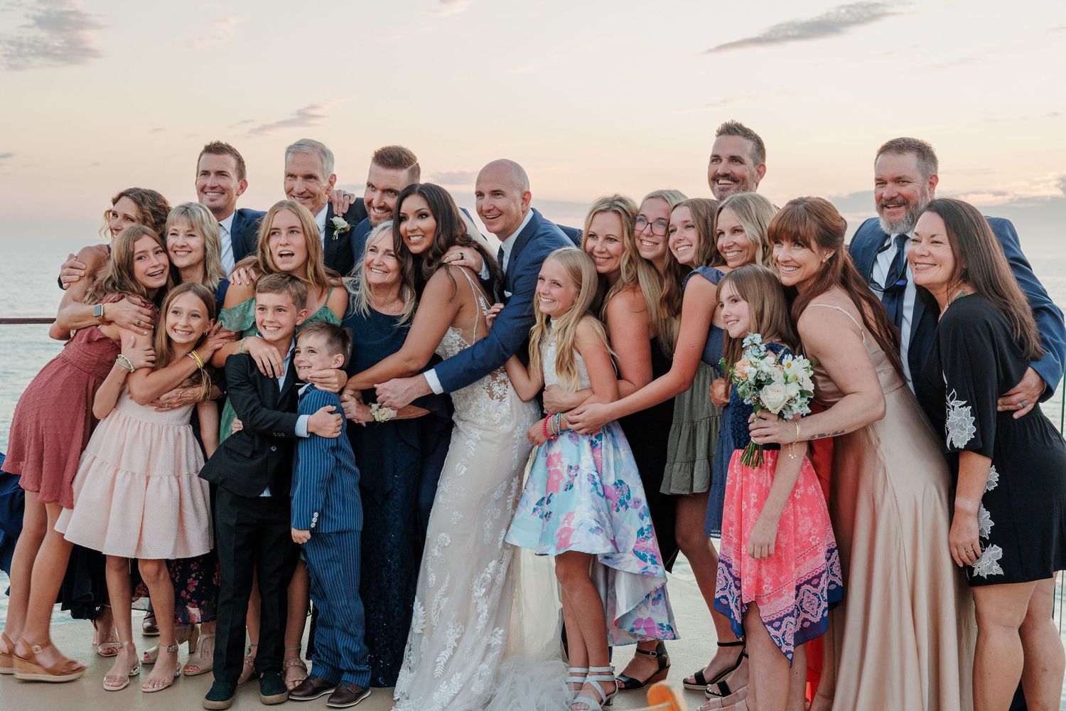 Large wedding party group photo with bride and groom surrounded by family at ocean sunset.