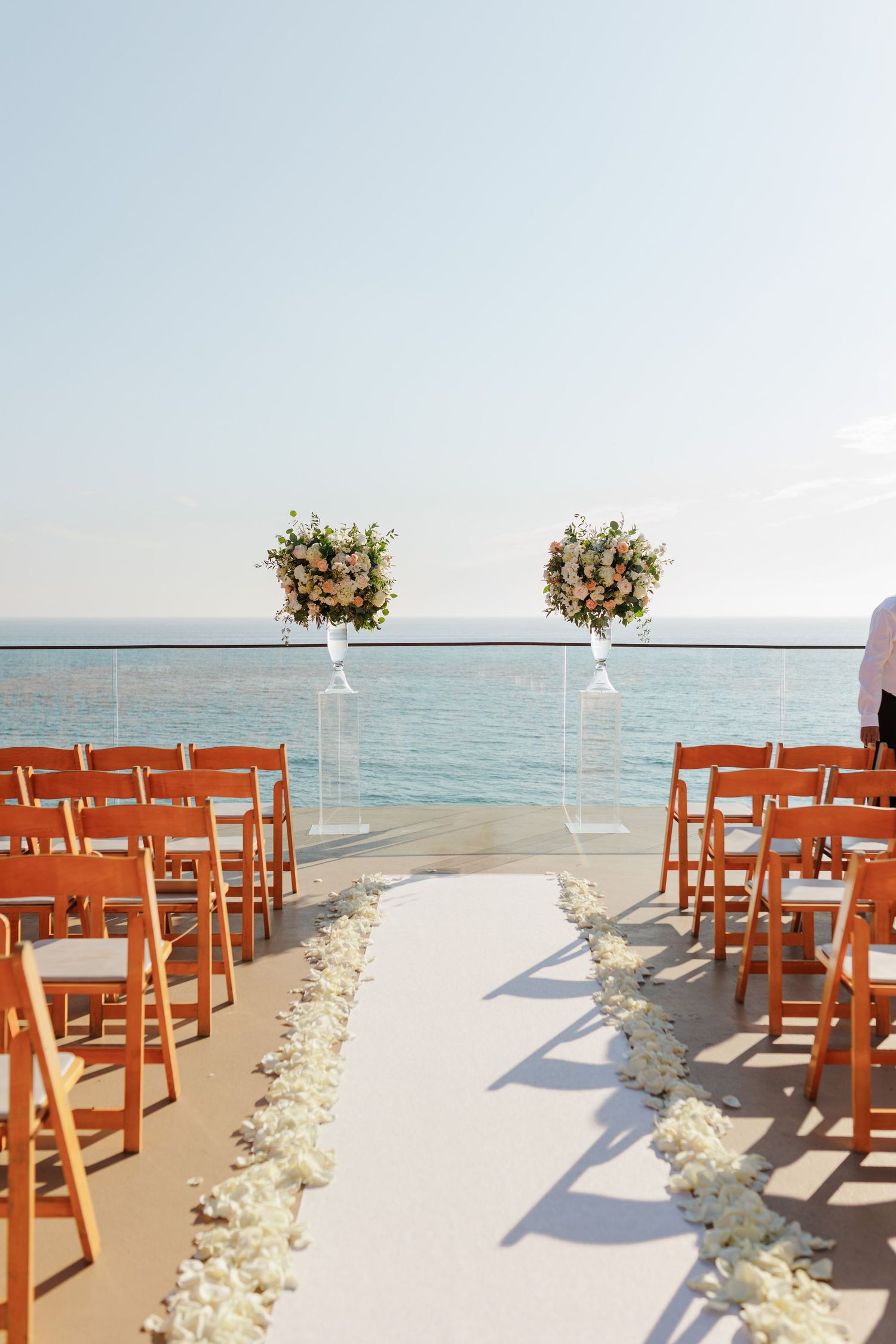 Elegant outdoor wedding ceremony setup with wooden chairs, floral arrangements, and ocean backdrop.