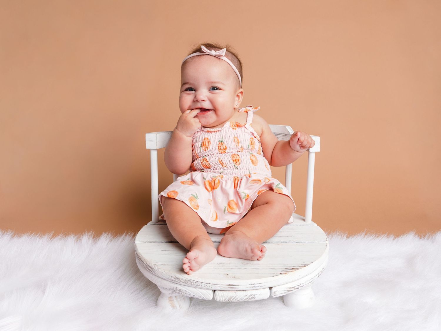 Smiling baby girl in orange floral romper sitting on white chair against tan background on fluffy white rug.