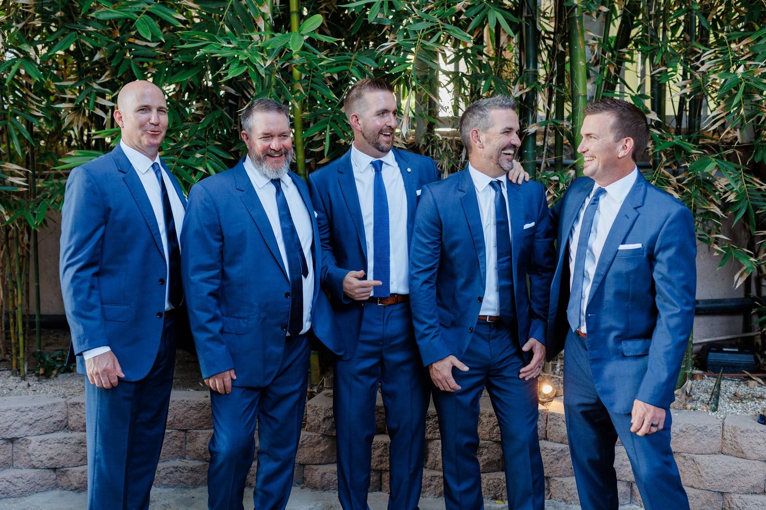 Five men in matching blue suits and ties laughing together outdoors at a wedding celebration.