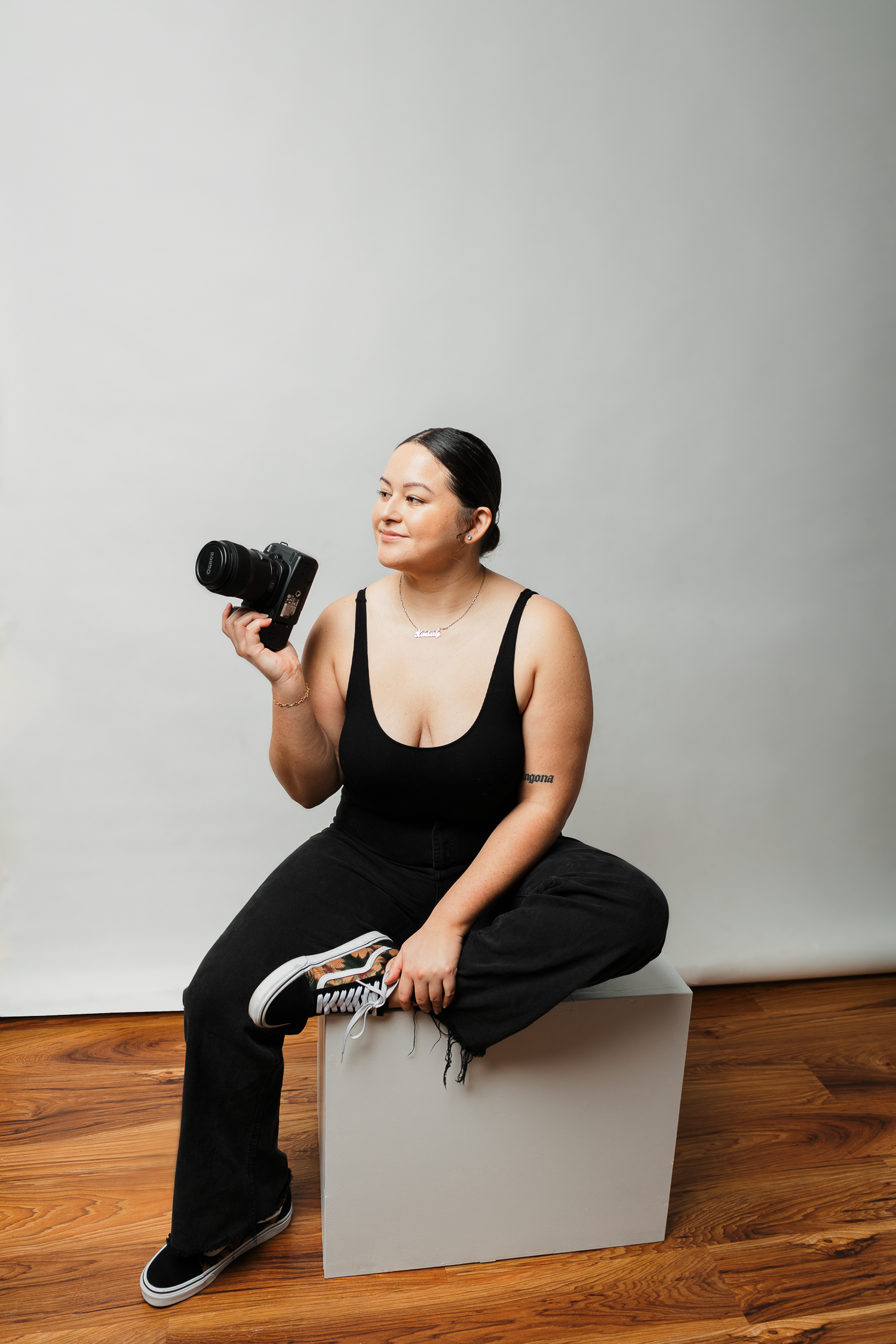 Photographer in black dress poses with camera against white backdrop in studio setting.