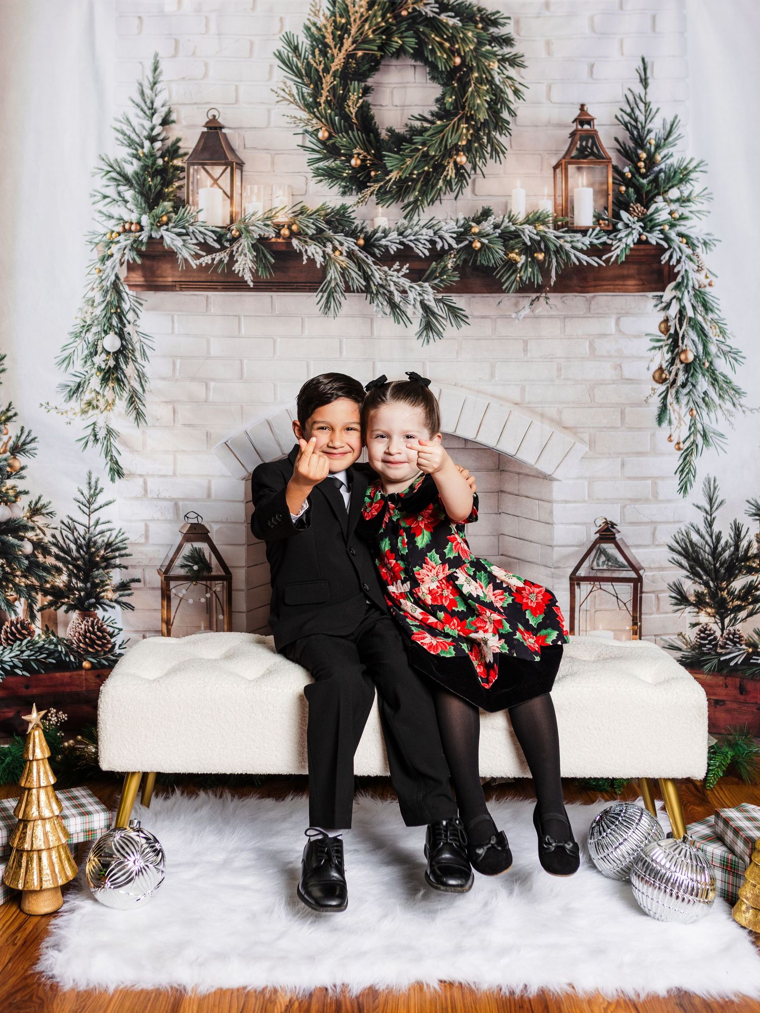 Two children in holiday attire pose on a bench before a decorated fireplace with wreaths and Christmas trees.
